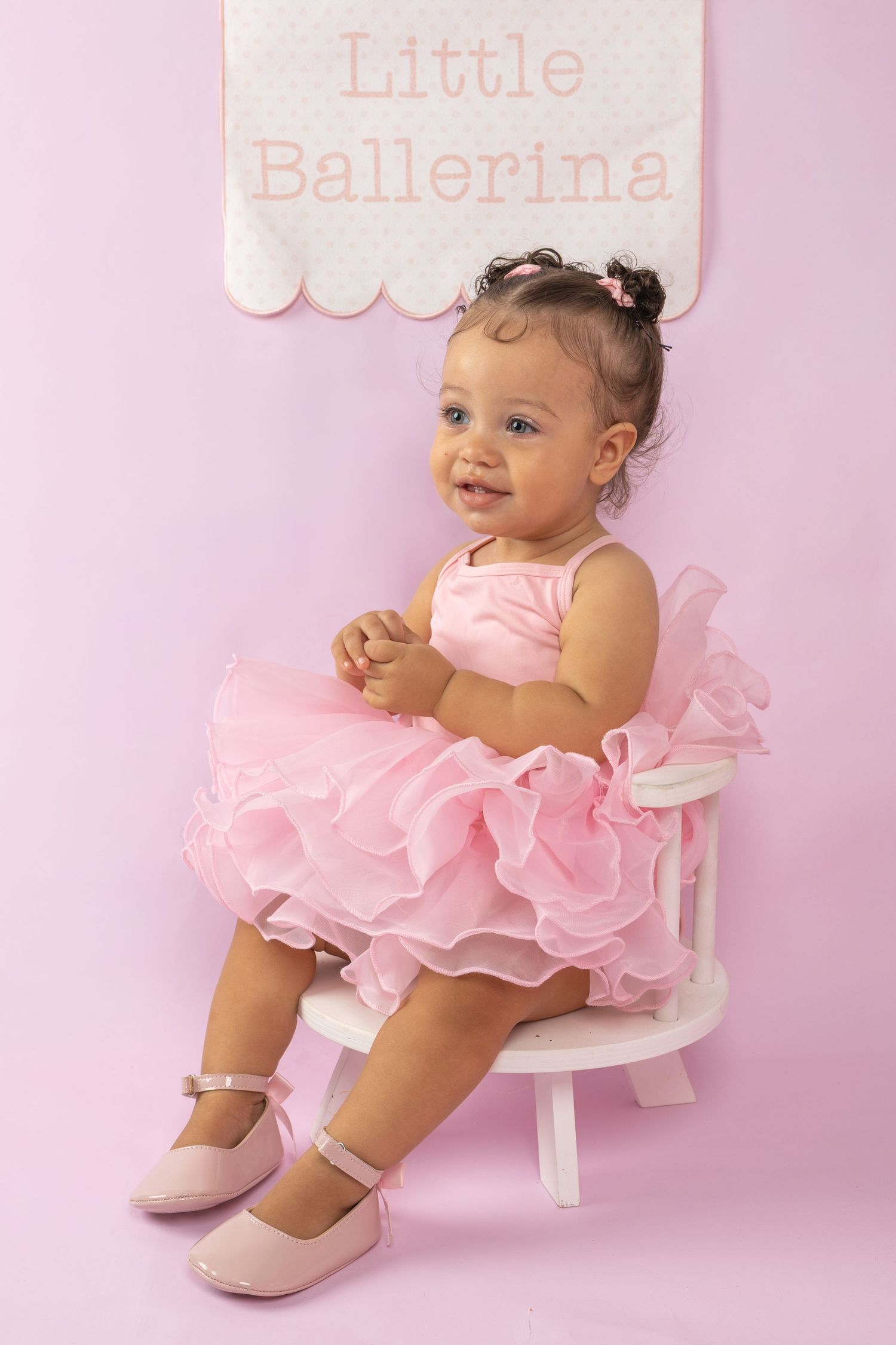 Smiling baby girl in pink tutu dress and shoes sits on white chair under Little Ballerina sign.