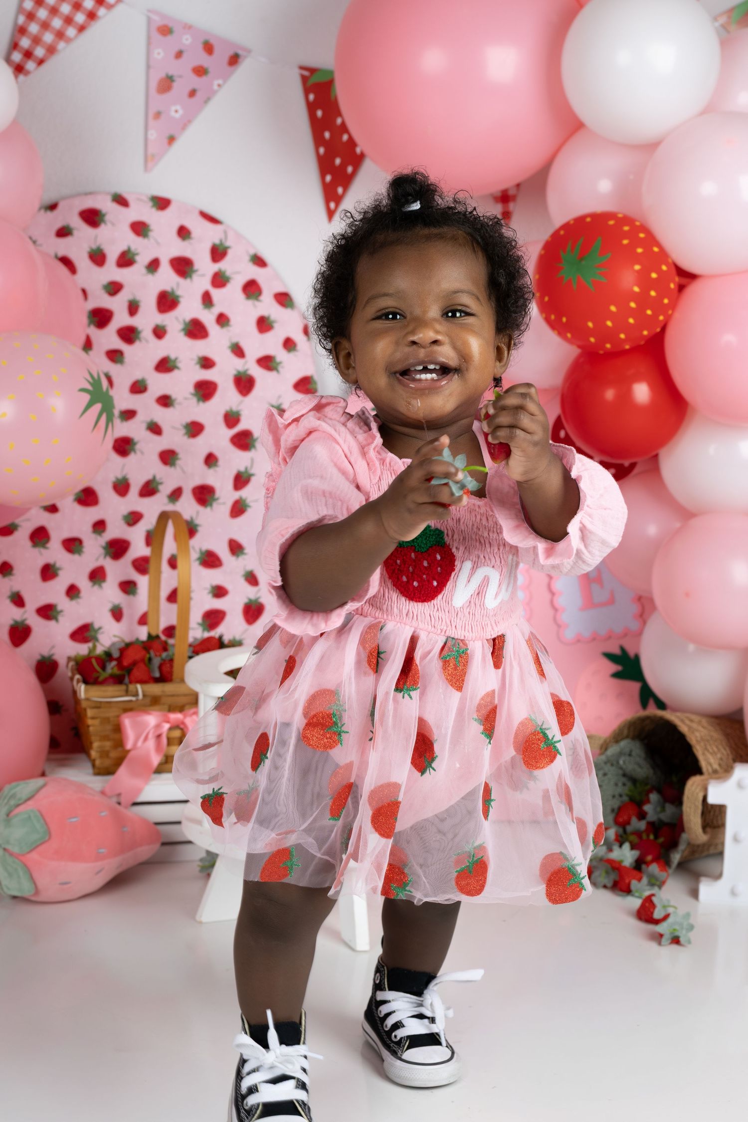 Smiling toddler girl in strawberry dress and Converse shoes at a pink strawberry-themed first birthday party.