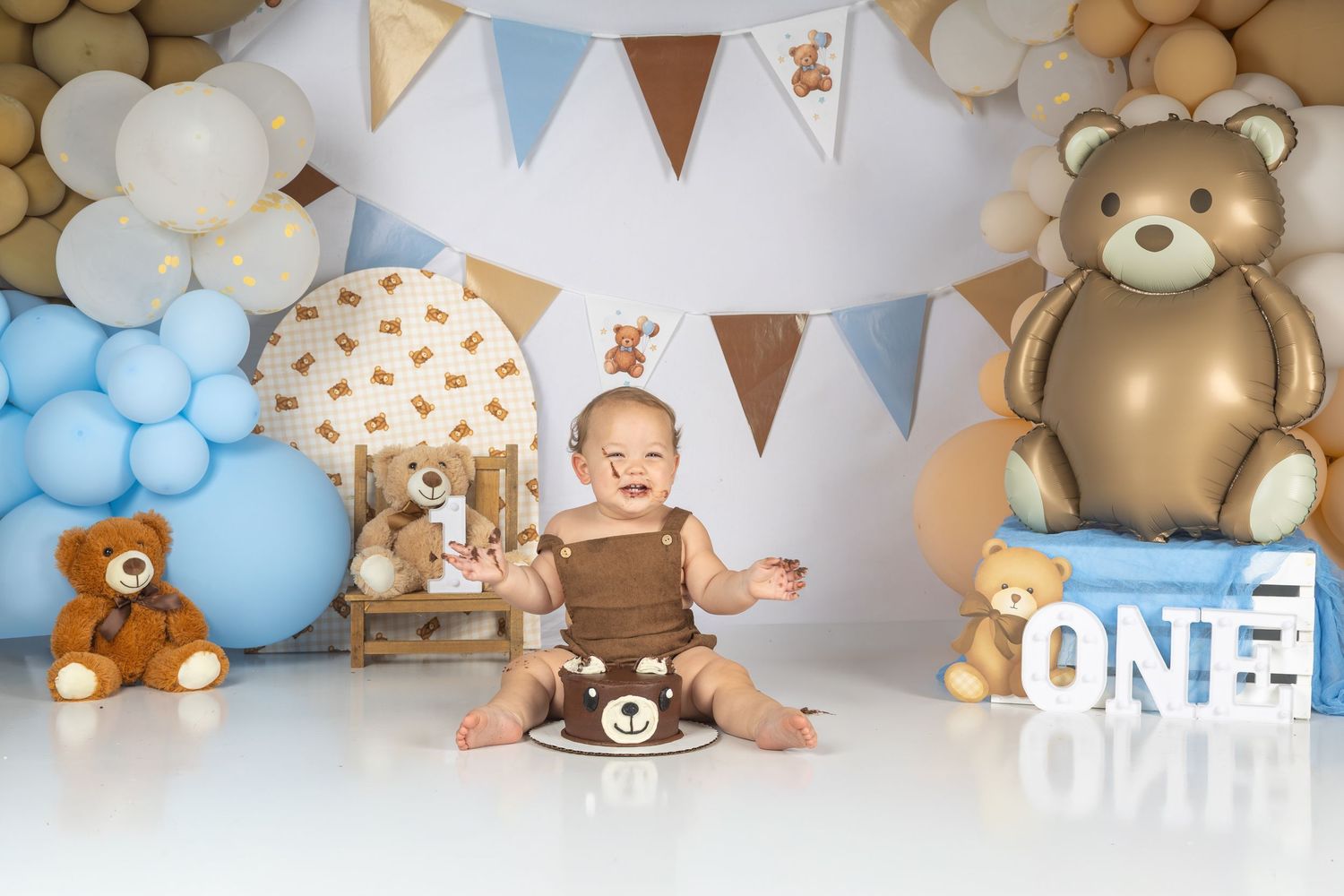 Baby's first birthday cake smash with teddy bear theme, blue and brown balloons, bunting flags, and ONE sign.