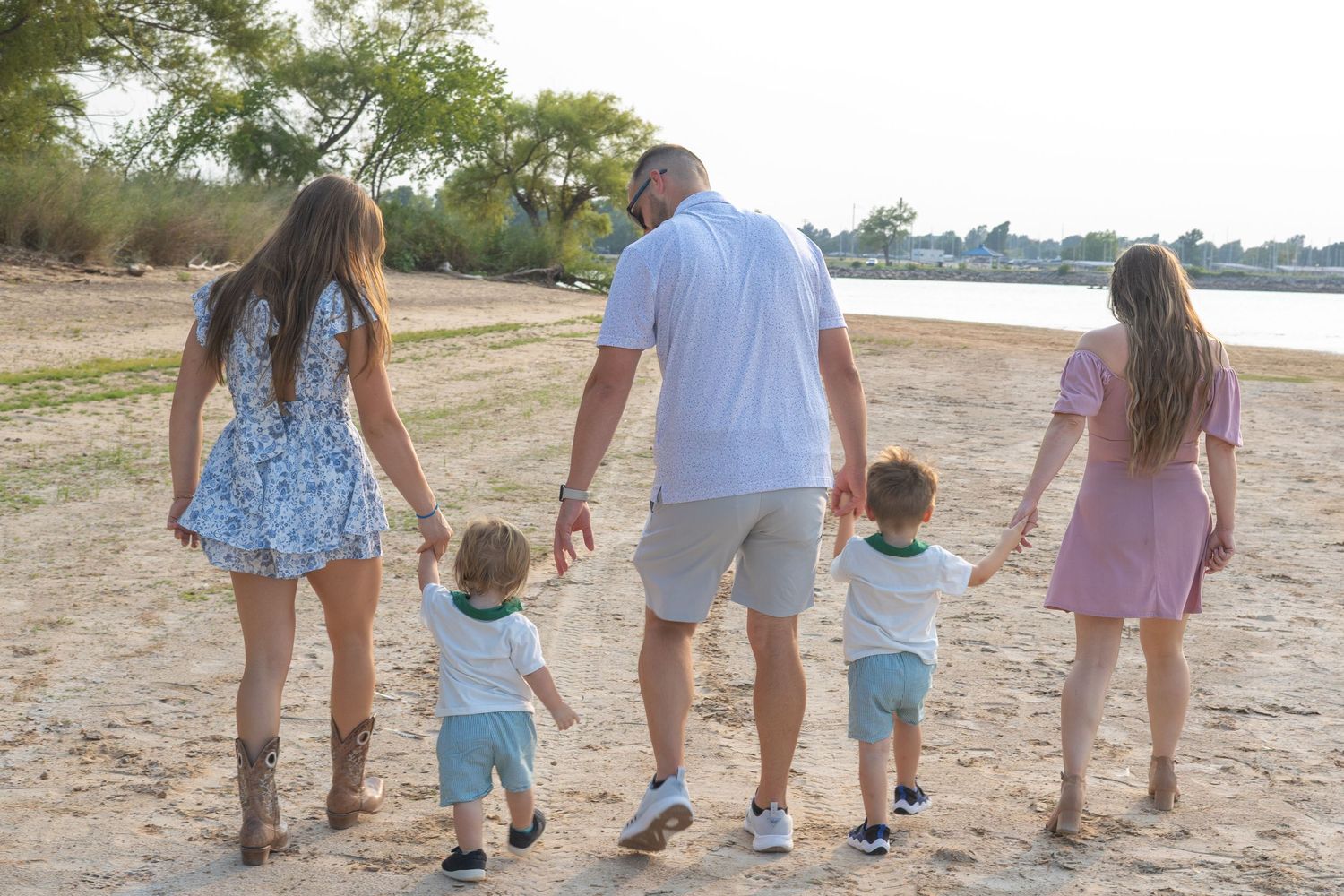 Family of five walking hand-in-hand on a sandy beach at sunset, with two toddlers between parents and older child.