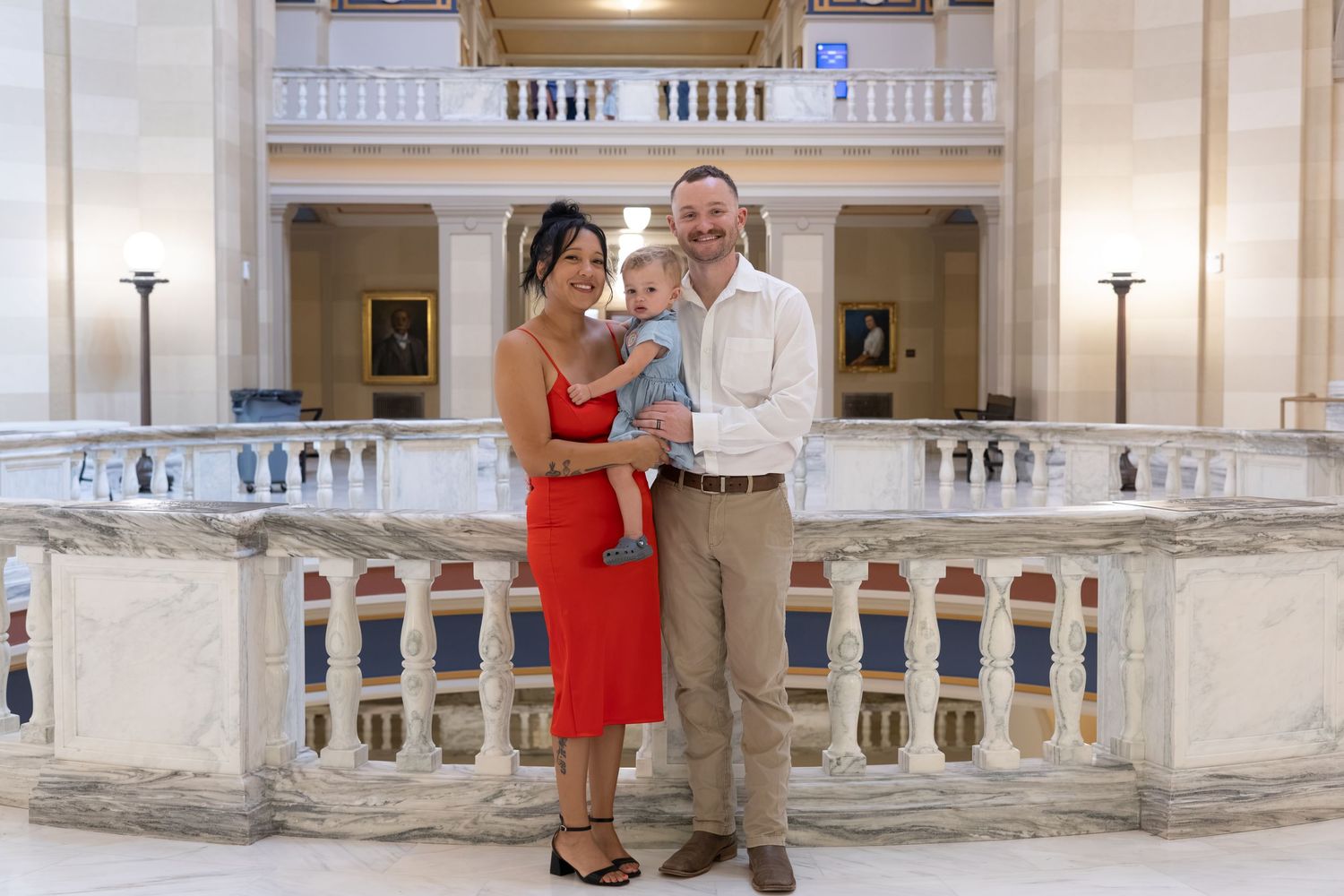 Family of three posing in elegant marble building interior, woman in red dress, man holding baby.