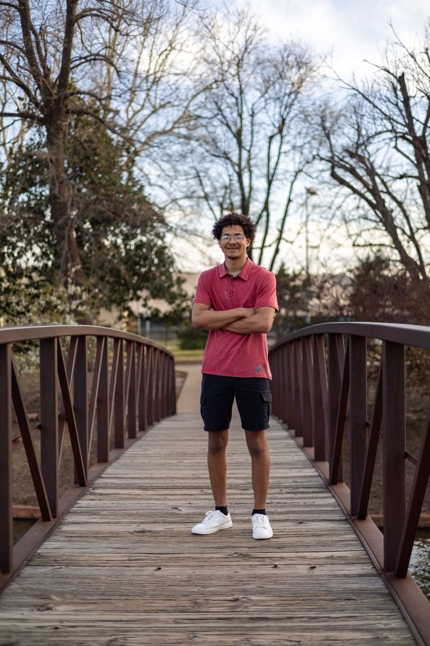 A person in pink shirt stands on a wooden footbridge in a park setting during dusk hours.