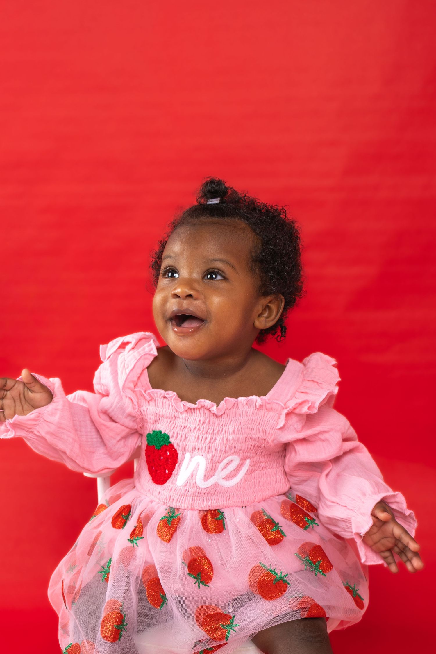 Smiling baby girl in pink strawberry dress celebrating her first birthday against a red background.