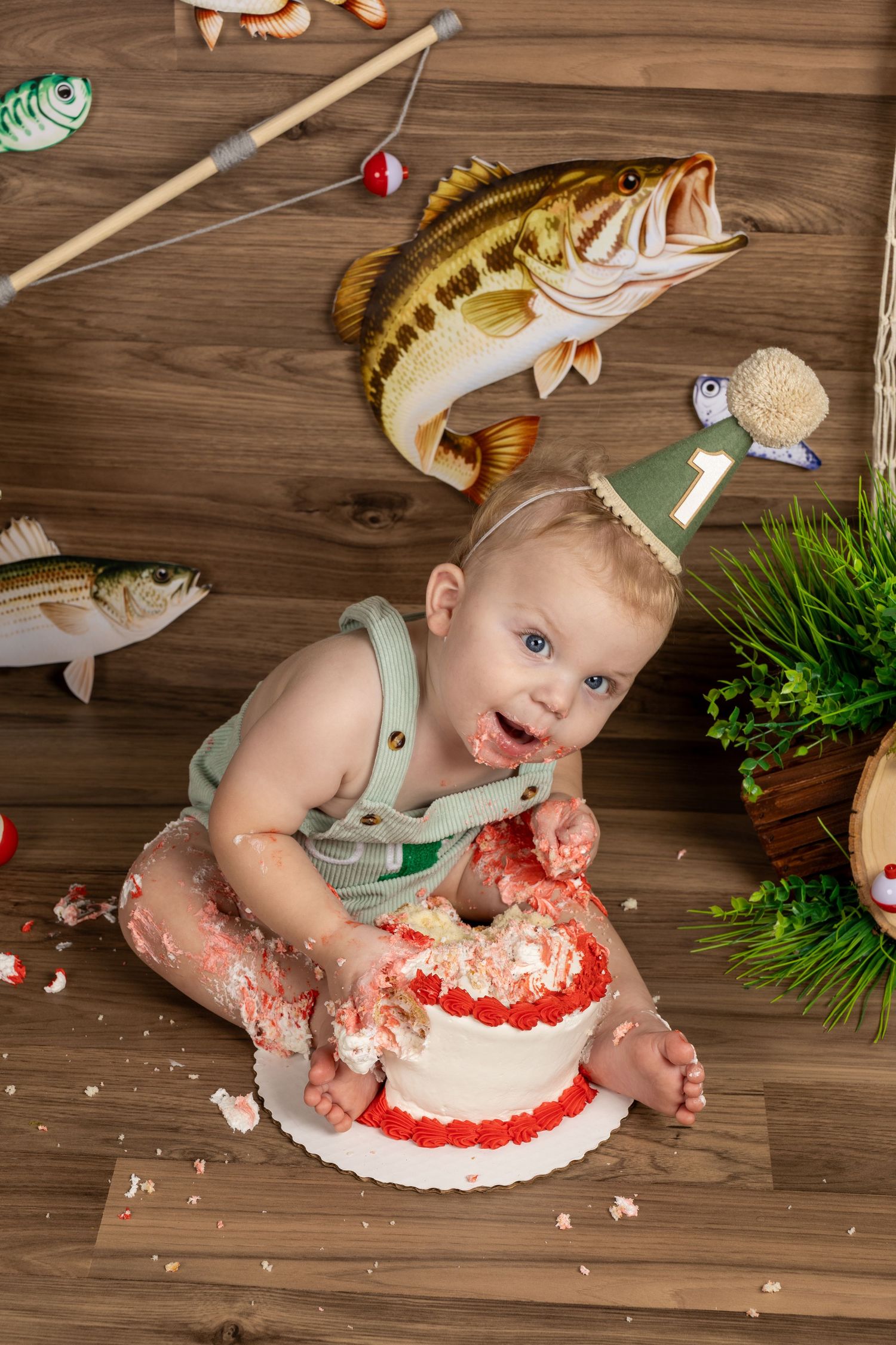 Baby in green overalls smashes first birthday cake at fishing-themed party with decorative bass fish and rod.