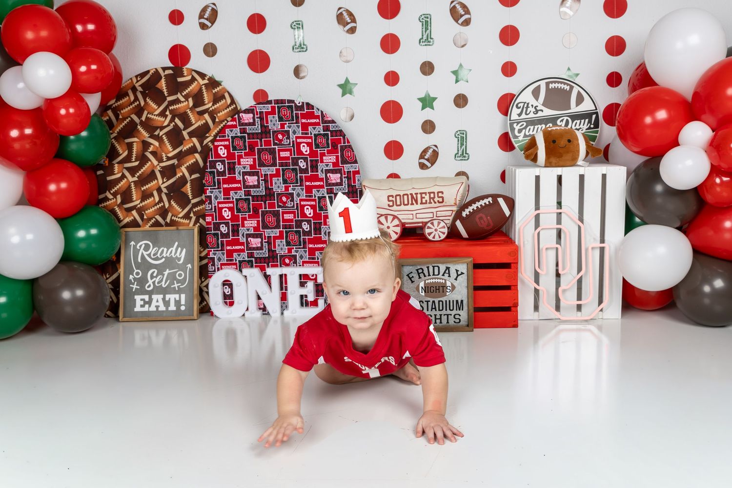 Baby boy in red outfit crawls at a sports-themed first birthday party with balloons and football decorations.
