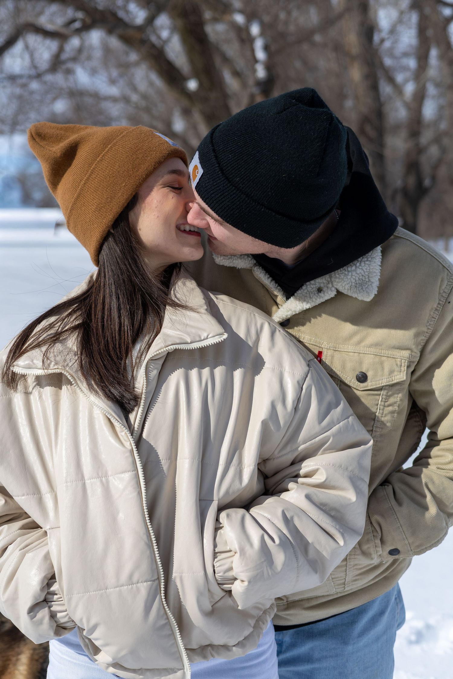 A couple in winter attire embraces in the snow, wearing beige jackets and beanies against a snowy landscape.