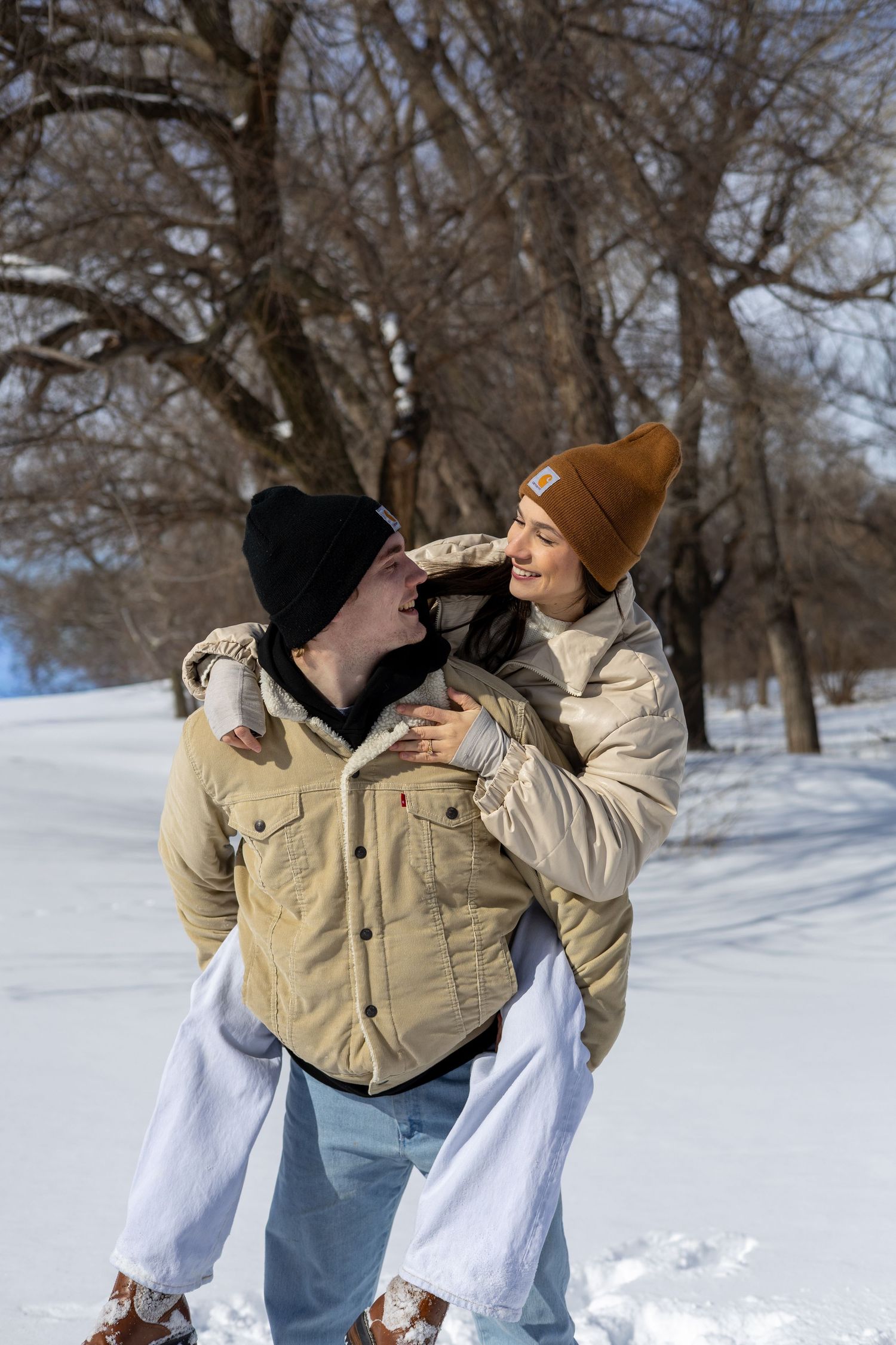 A couple in winter coats share playful moments in the snow, with bare trees visible in the background.
