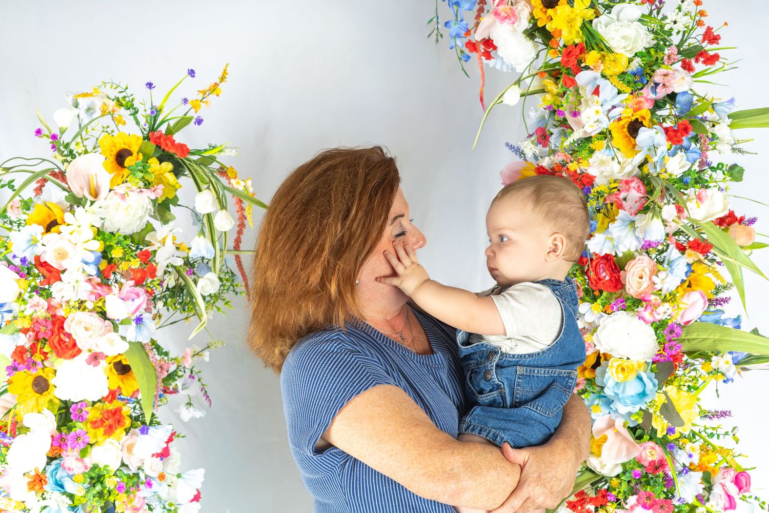 Woman with red hair holds smiling baby in denim overalls surrounded by colorful floral arrangements.