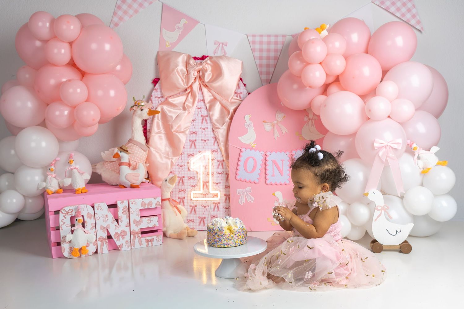 Baby girl in pink dress celebrates first birthday with ONE letters, pink balloons, and smash cake in a decorated studio.