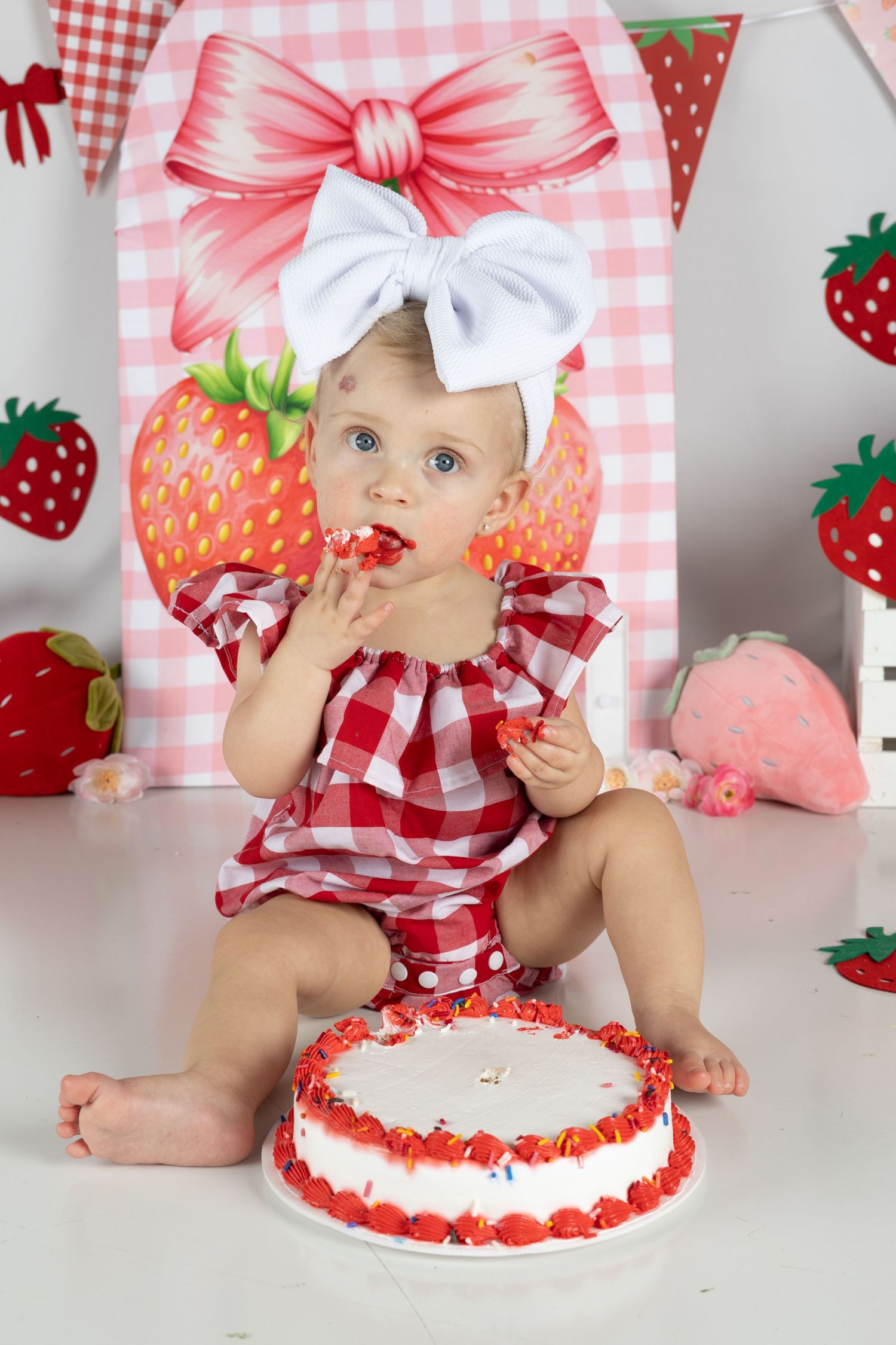 Baby girl in red gingham dress eating cake at strawberry-themed first birthday cake smash photo session.