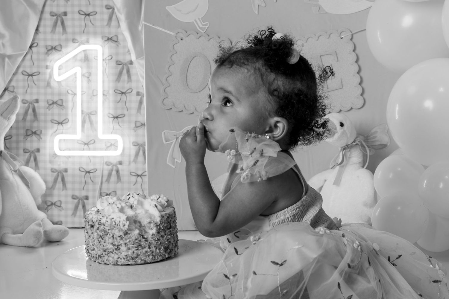 Black and white photo of a baby girl celebrating her first birthday with cake and a neon number one sign.