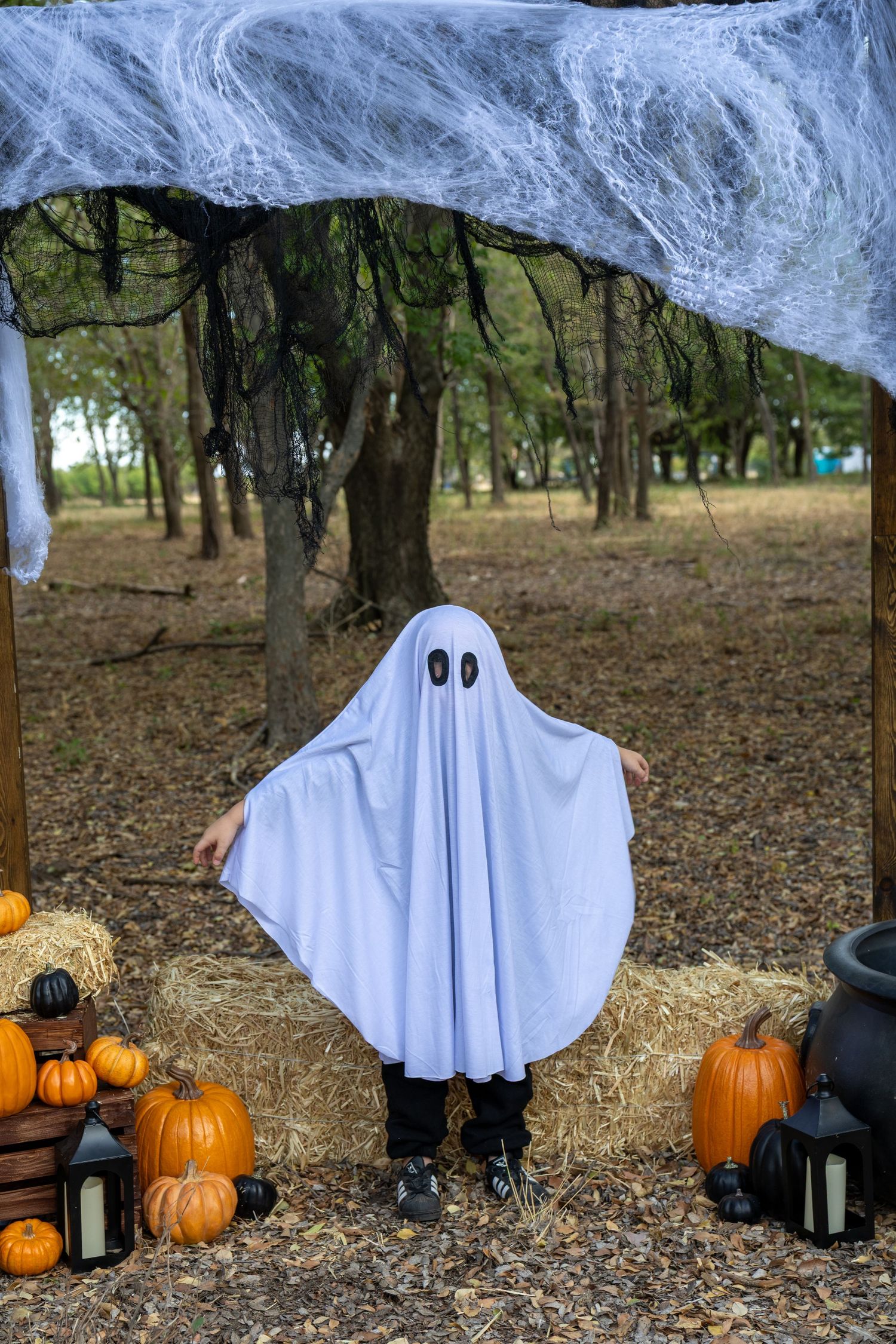 Child in white ghost costume poses with pumpkins and hay bales under spooky Halloween decorations outdoors.