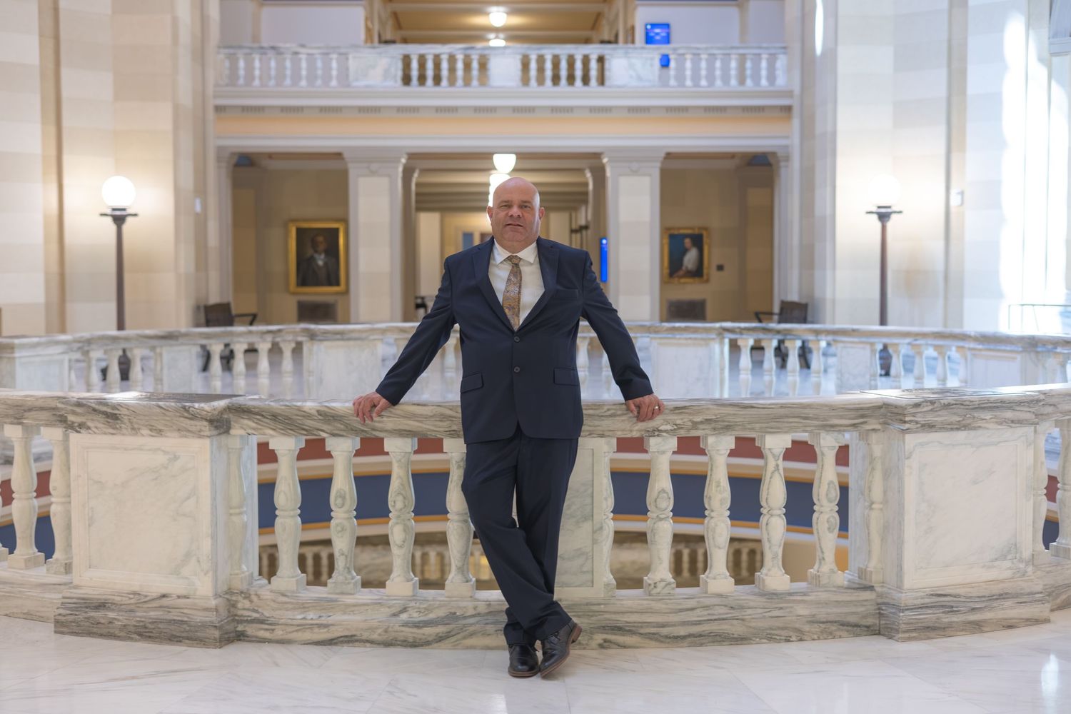 Man in navy suit posing confidently in elegant marble government building interior with ornate balustrades.