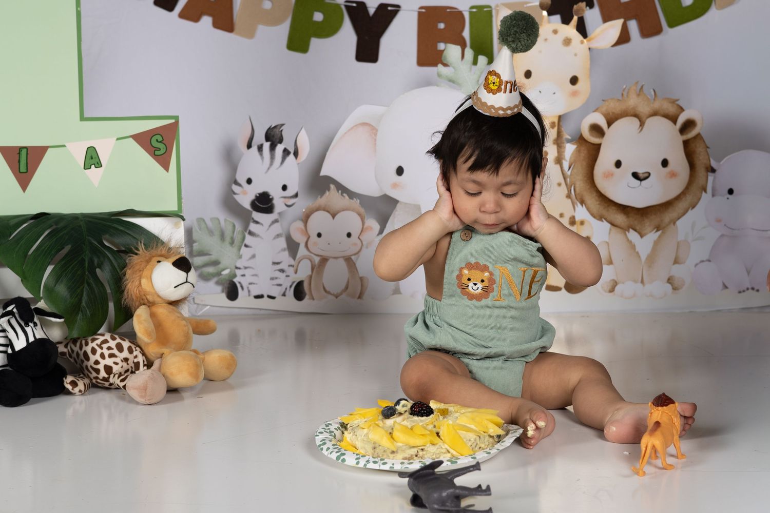 Baby in safari-themed first birthday setup with animal toys, cake smash, and Happy Birthday banner.