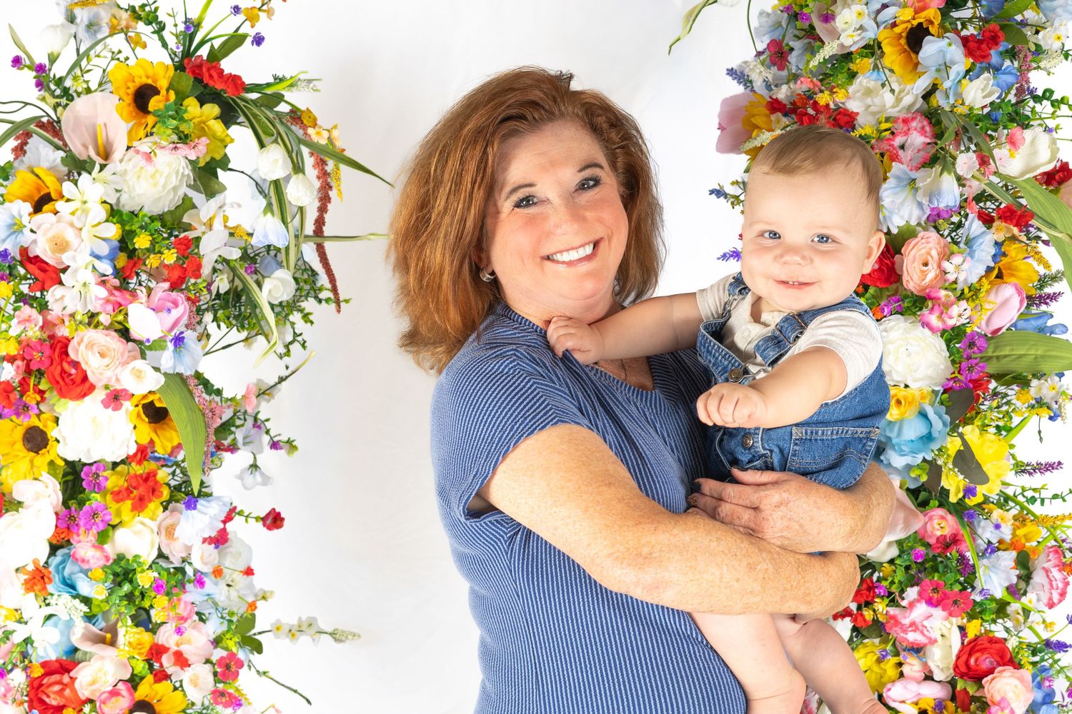 Woman holding a baby surrounded by colorful floral arrangements in a bright portrait setting.