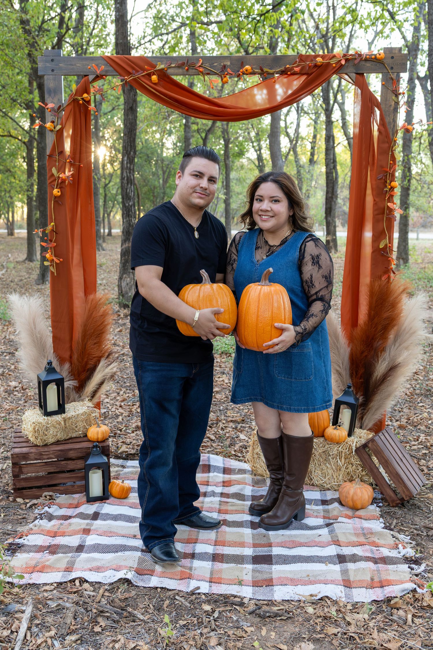 Couple holding pumpkins in front of a rustic fall arch with orange draping, hay bales, and lanterns in an autumn forest.