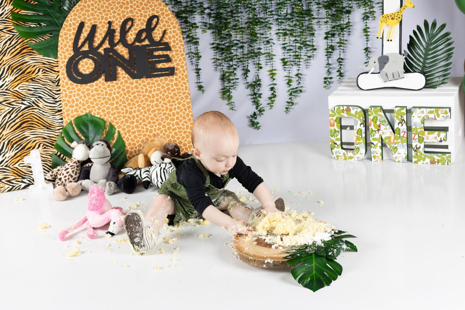 Baby in black outfit smashing first birthday cake at Wild One jungle themed party with tropical decorations.