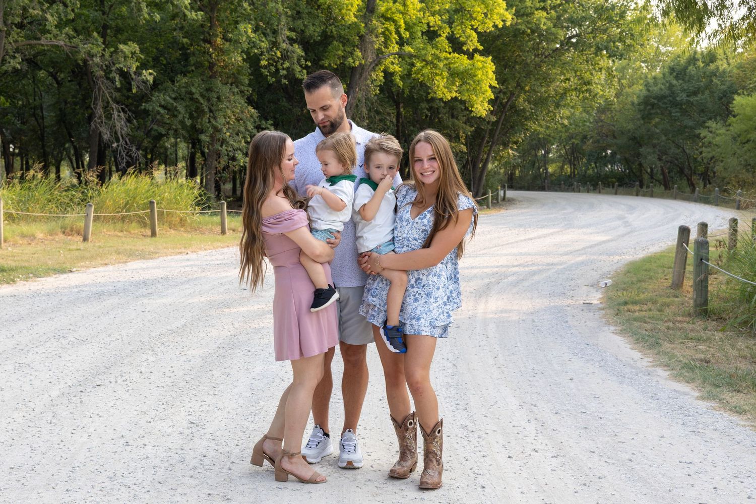Family of five posing on a gravel country road surrounded by lush green trees on a sunny day.