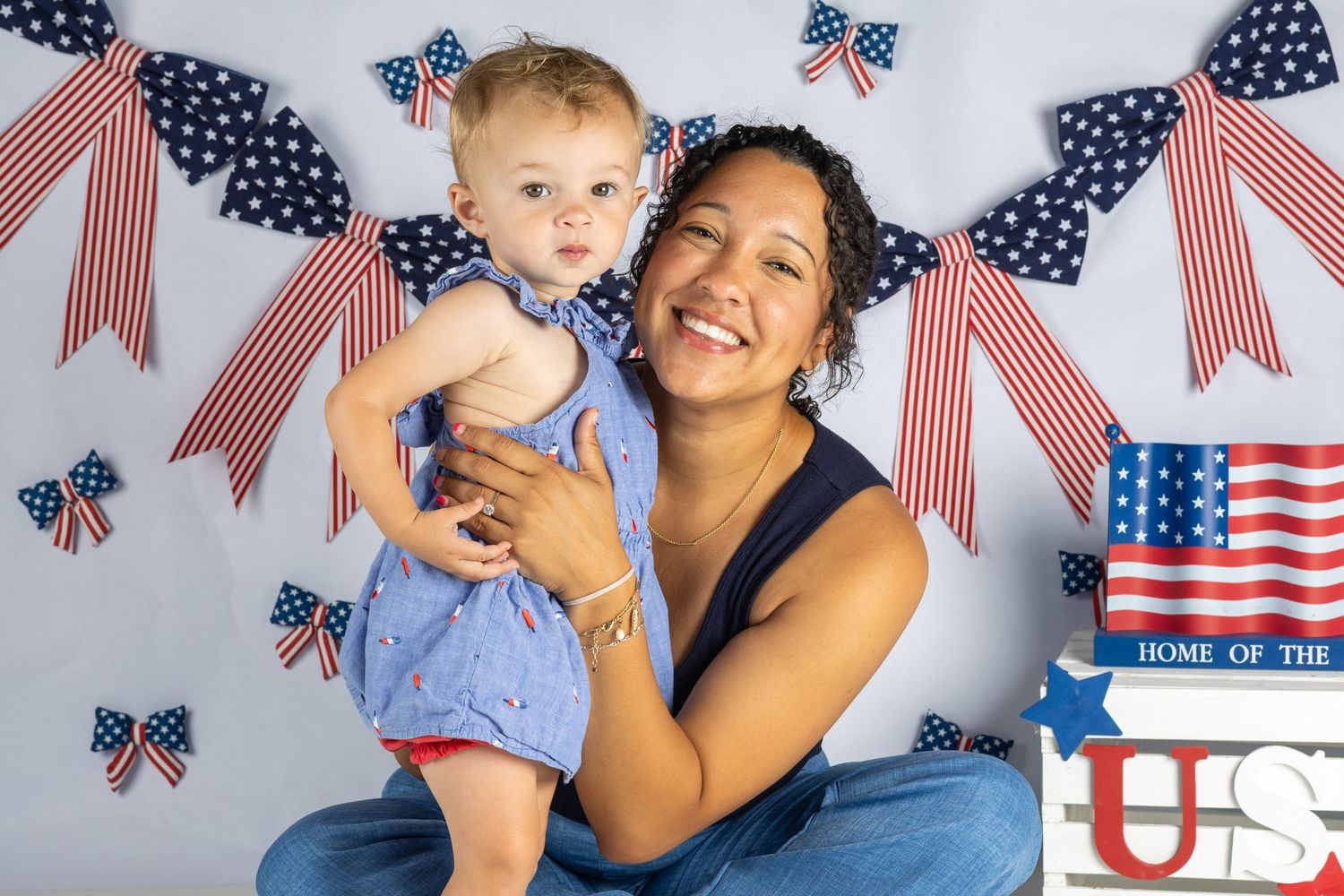 Woman and baby in patriotic outfits pose with American flag bows and US decorations for a 4th of July photo.