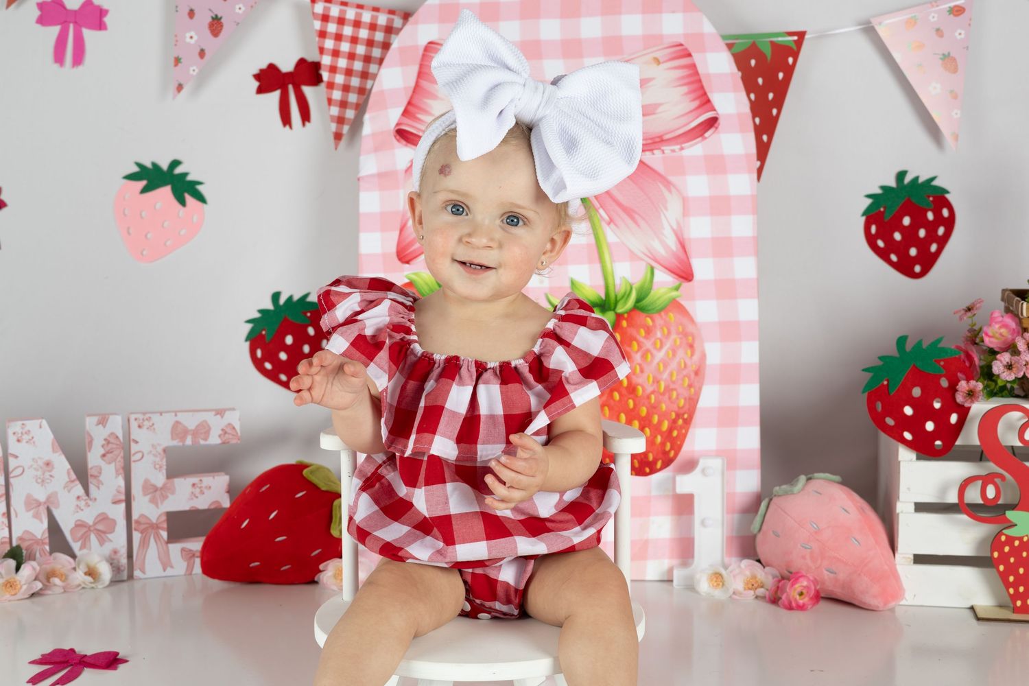 Baby girl in red gingham romper with white bow headband poses at strawberry-themed first birthday cake smash photo session.