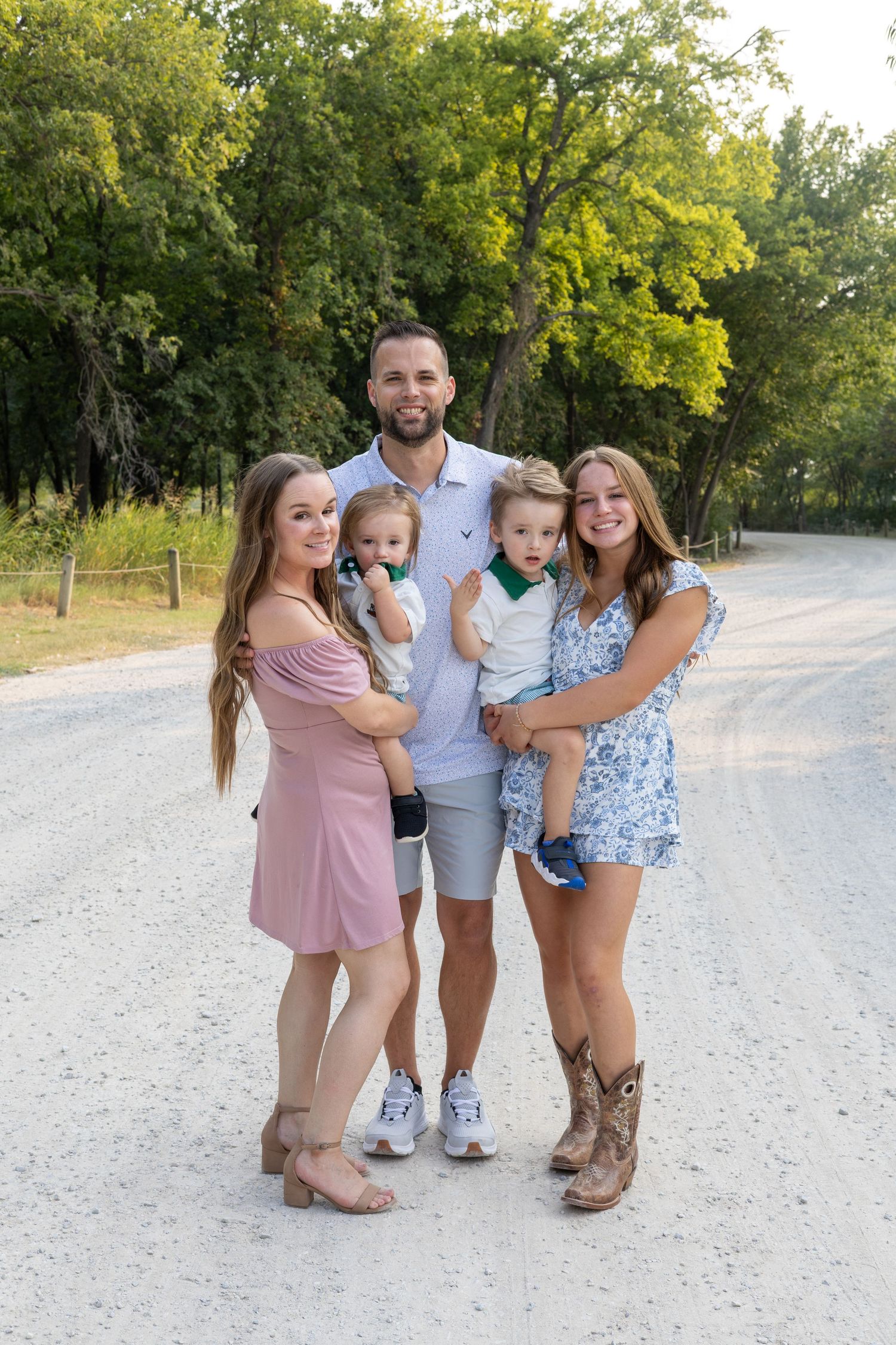 Happy family of five posing on a gravel path surrounded by lush green trees on a sunny day.