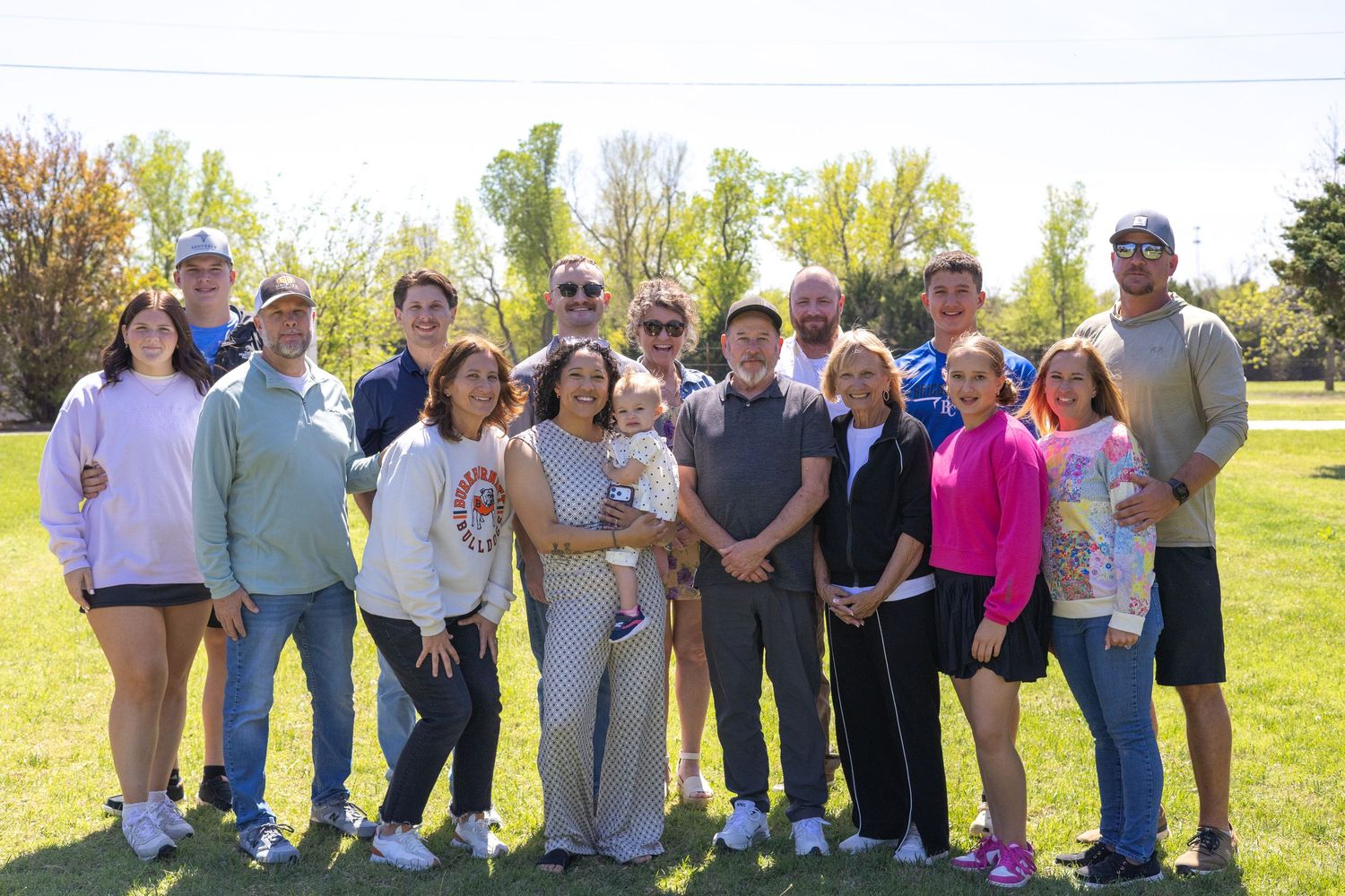 Large family group smiling outdoors on a sunny day in a green park with autumn trees in the background.