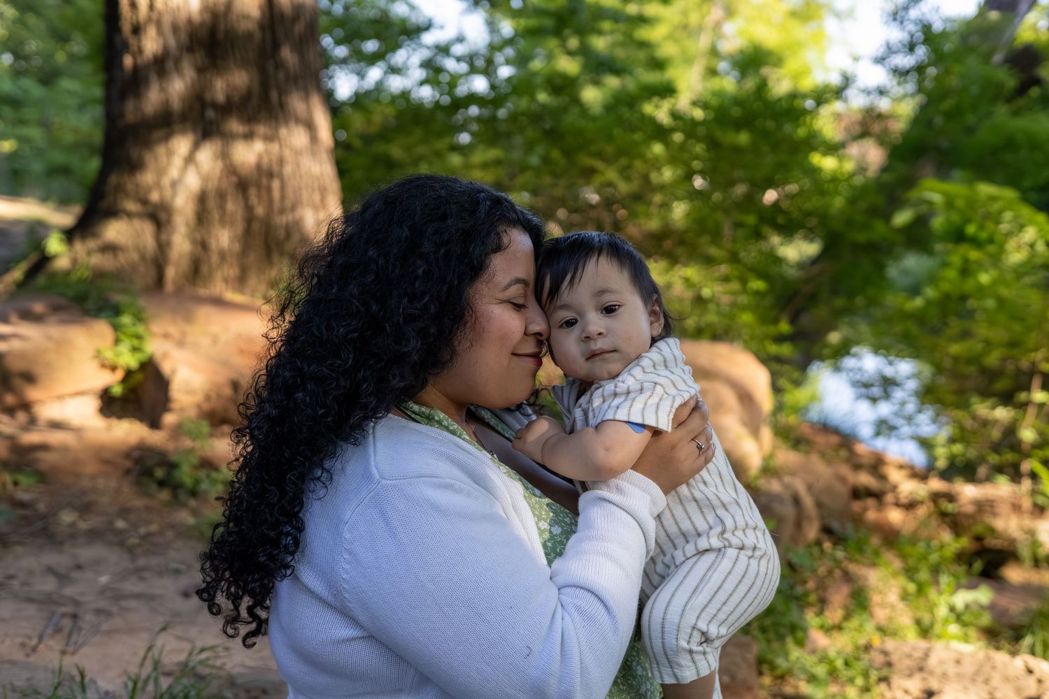 A mother with curly hair tenderly holds her baby outdoors in a lush green forest setting.