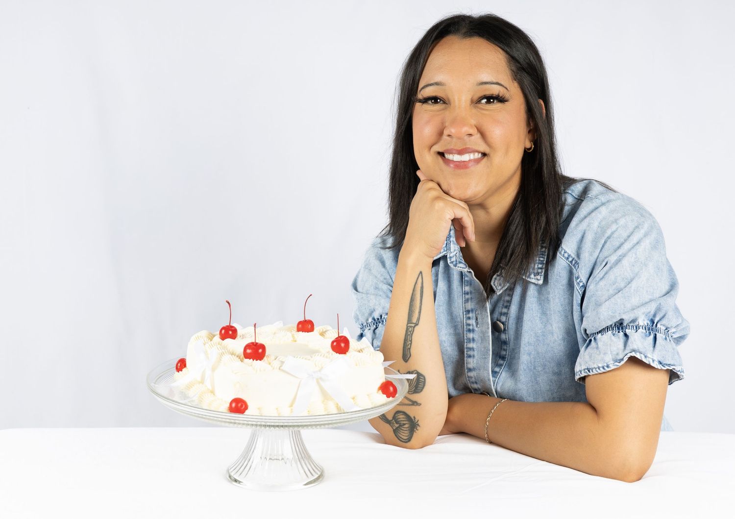 Woman smiling beside a white cake with cherries on a pedestal stand against a light background.