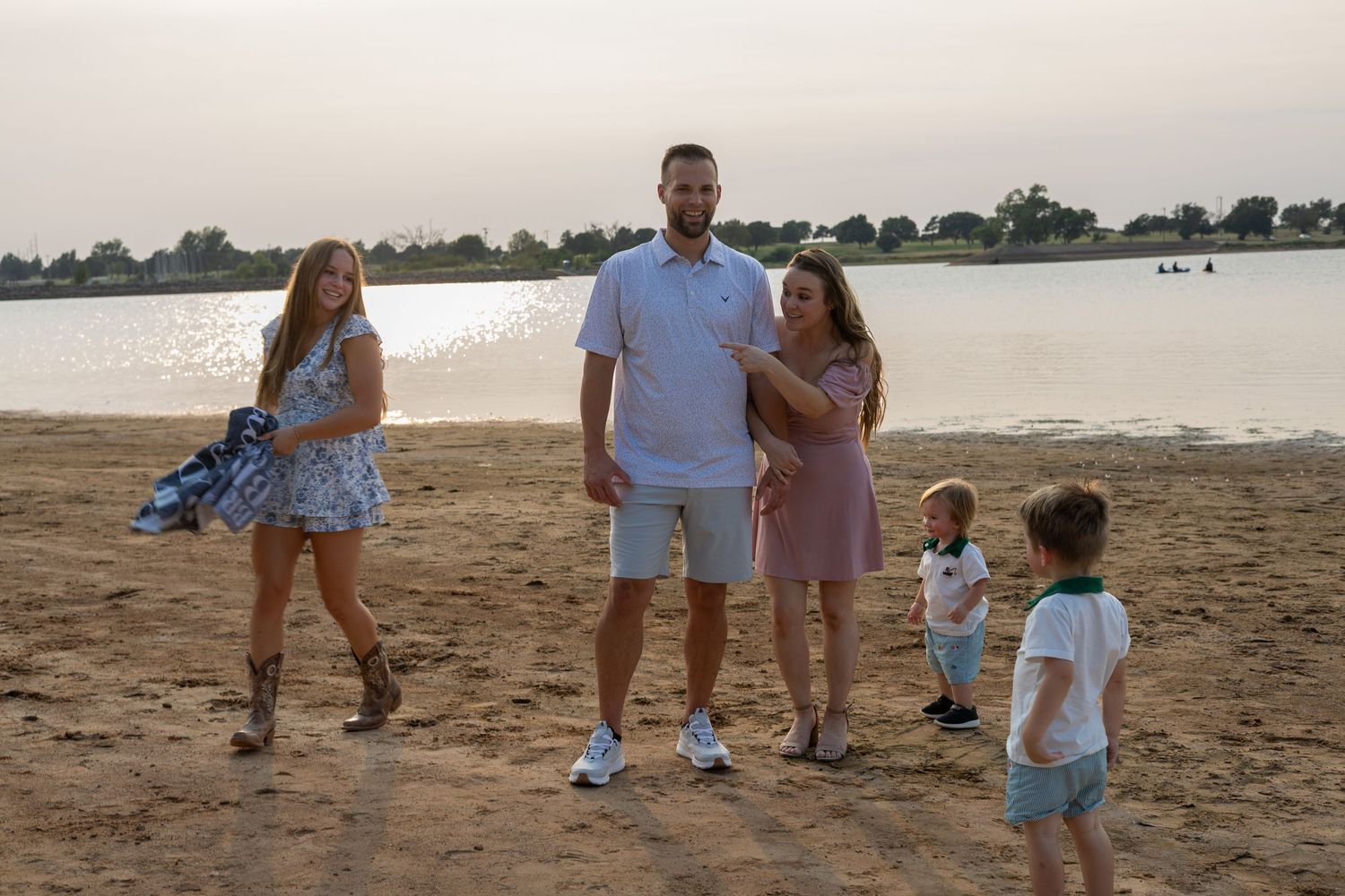 Family of five posing on sandy beach at sunset near calm lake, parents with three young children in casual summer outfits.