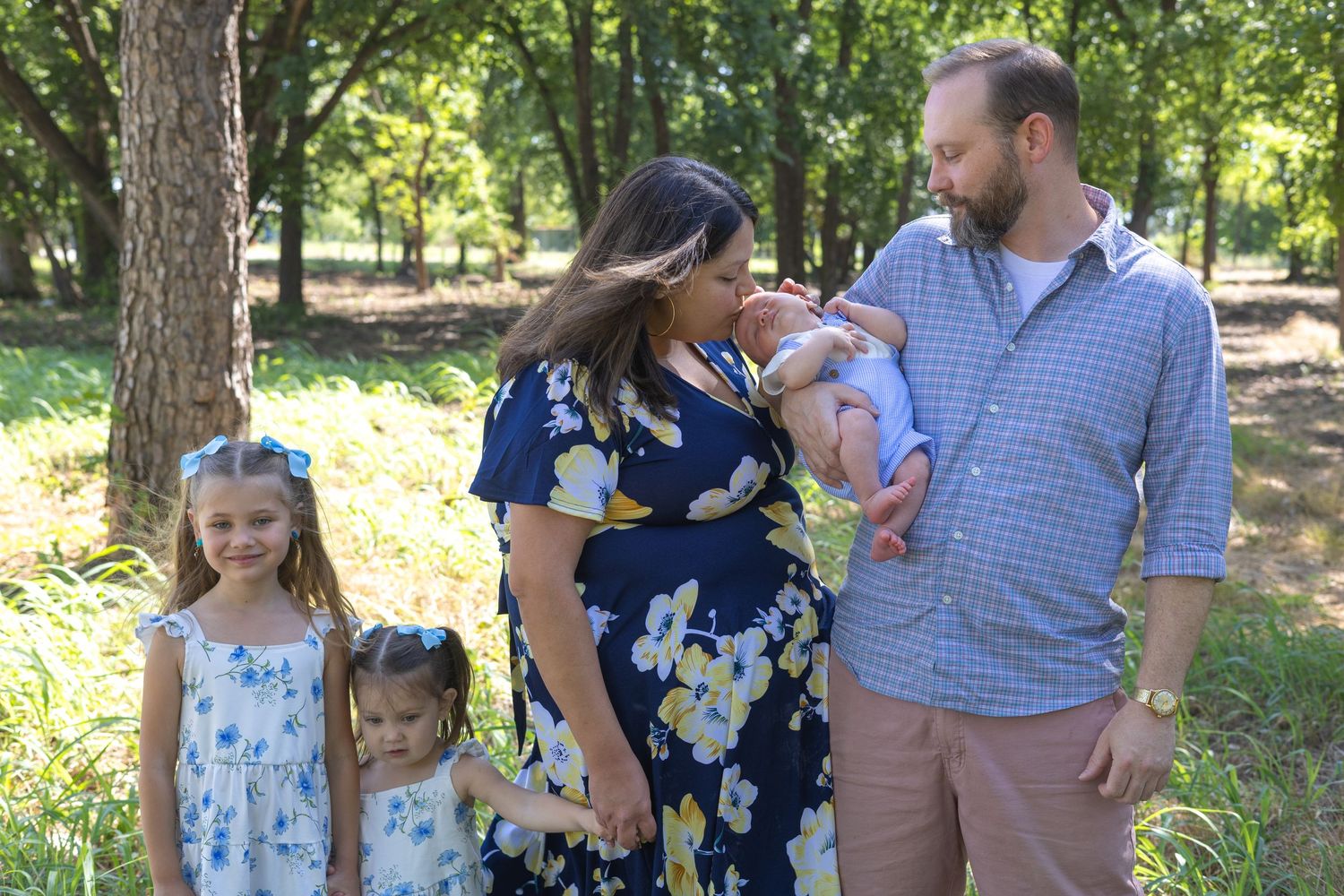 Family of five posing outdoors in a wooded park, with two girls, a toddler, newborn baby, and smiling parents.