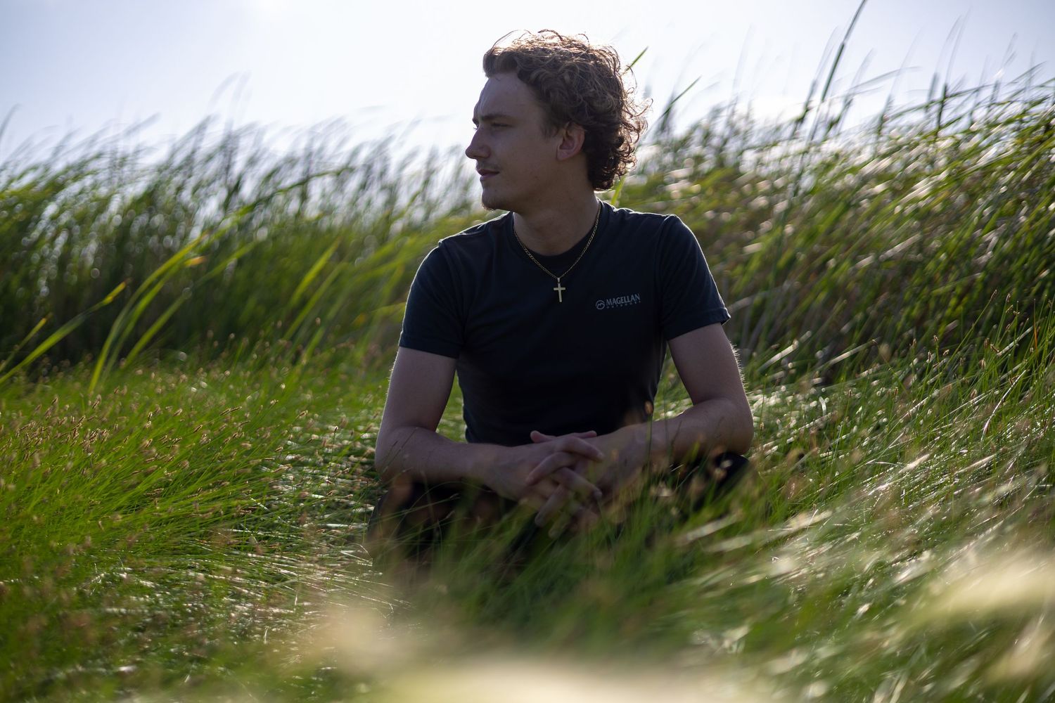 Young man in navy t-shirt sitting in tall green grass field, wearing cross necklace, looking thoughtfully into distance.