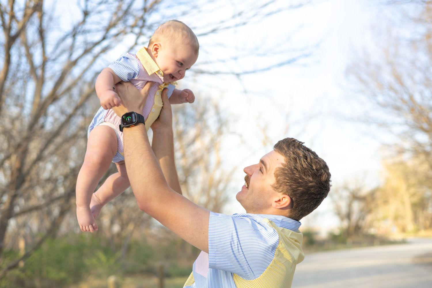A parent lifts a smiling baby wearing a diaper high into the air on a sunny day with bare trees in the background.