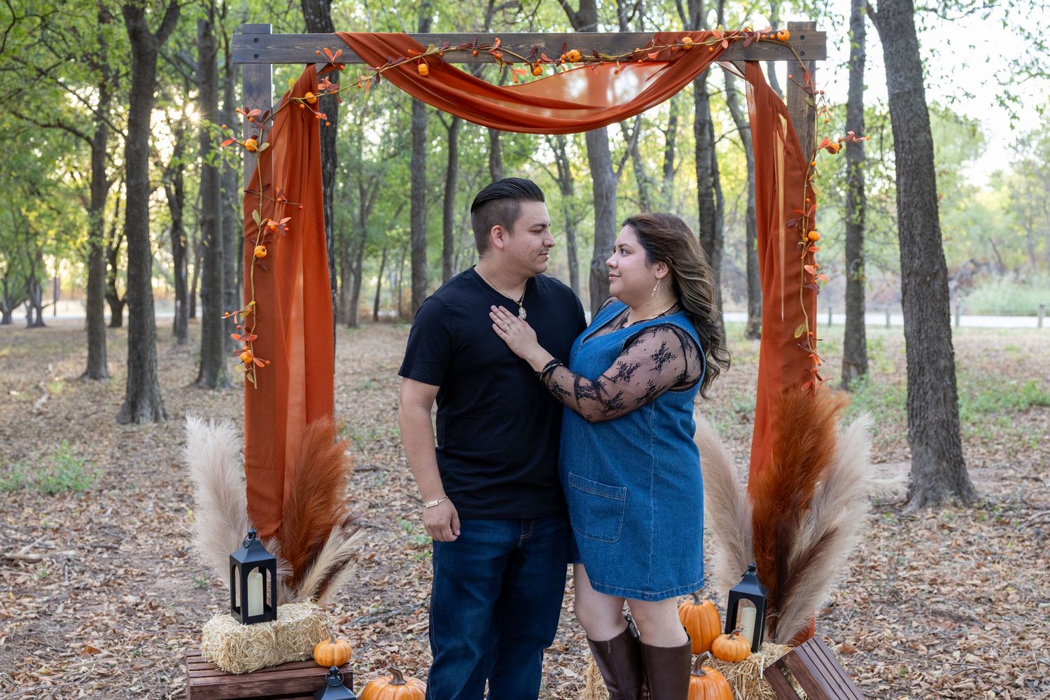 Couple embracing under rustic fall arch with pumpkins and orange draping in autumn forest setting.