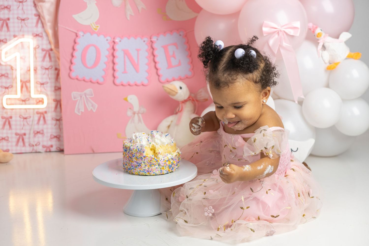 Baby girl in pink dress smiles at her first birthday smash cake surrounded by balloon decorations.
