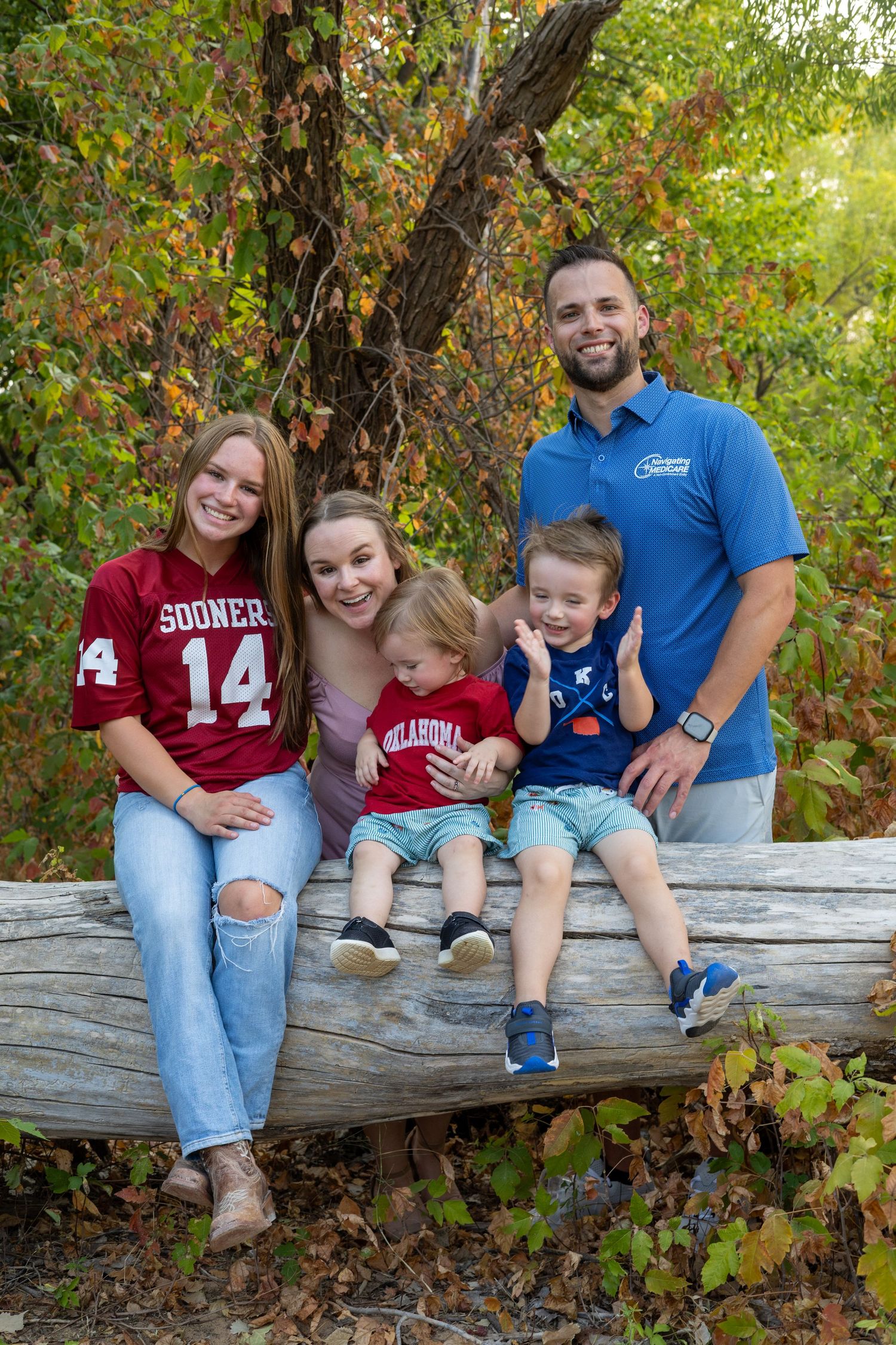 Family of five posing on a log in autumn, wearing Oklahoma Sooners gear, surrounded by colorful fall foliage.