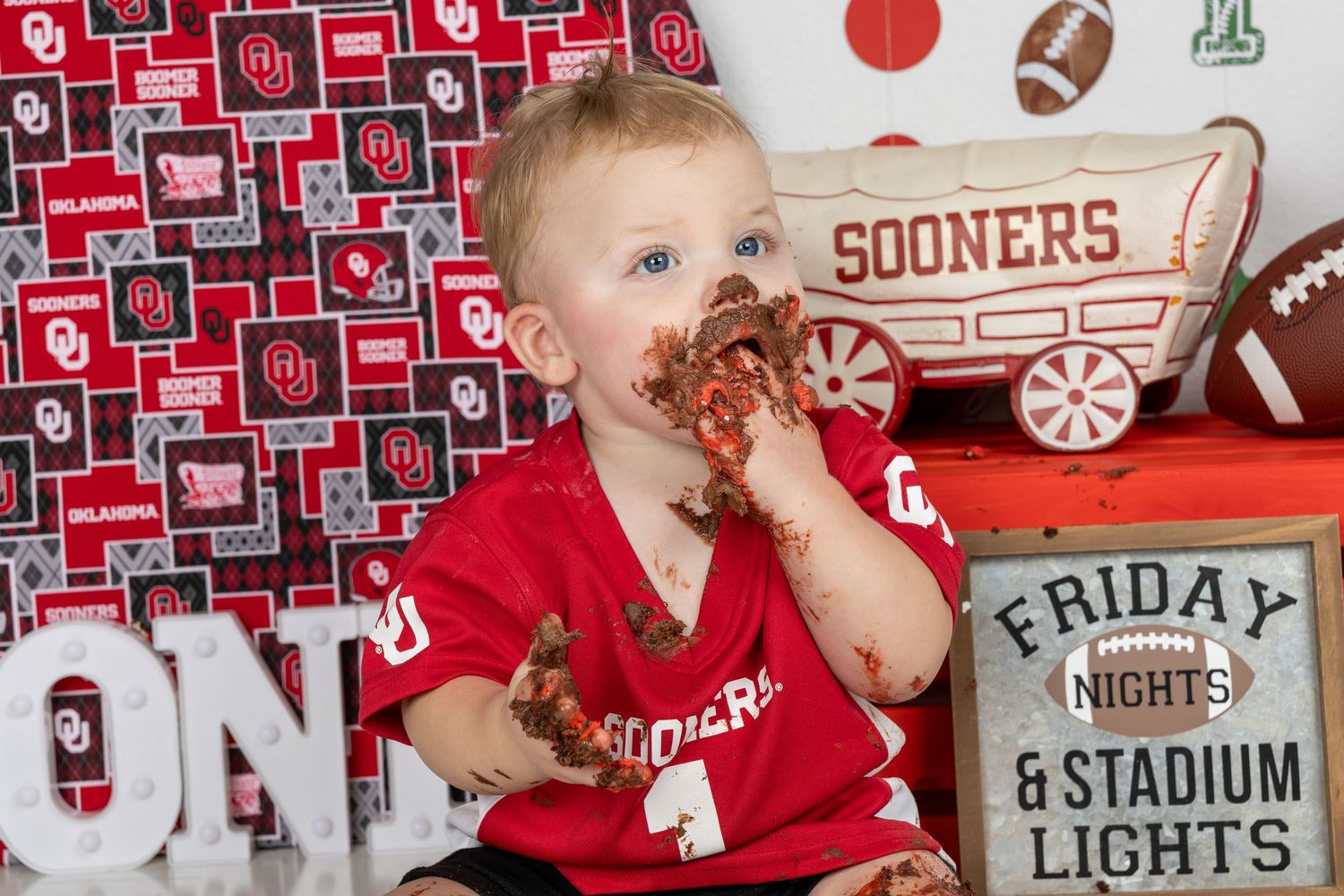 Baby in Oklahoma Sooners jersey enjoys smash cake surrounded by OU-themed decorations and a Friday Night Lights sign.