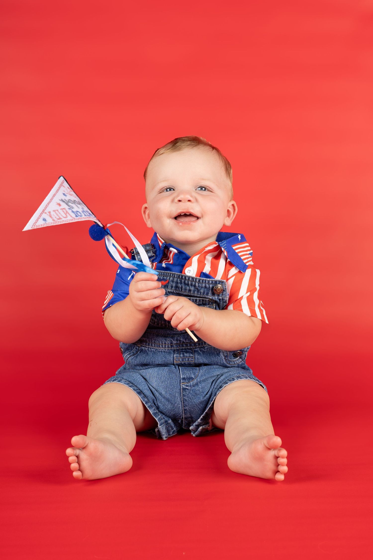 Happy baby in denim overalls and striped shirt holds patriotic flag toy, sitting on red background.