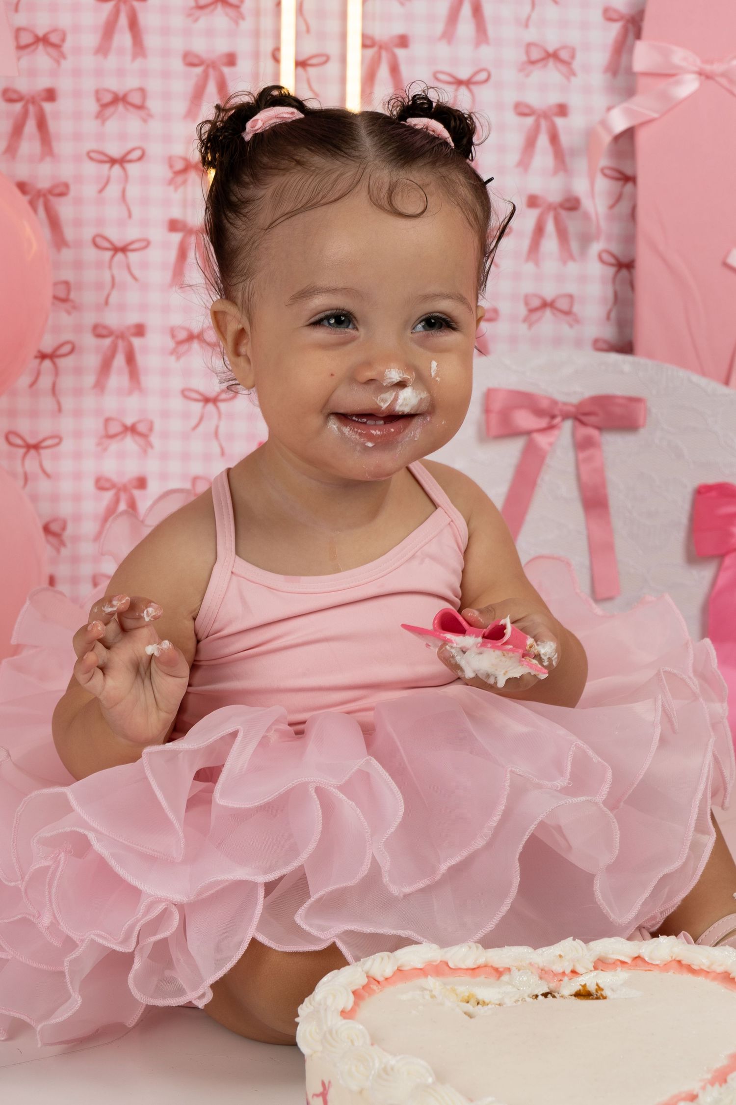 Baby girl in pink tutu smashes first birthday cake, face covered in frosting, surrounded by pink bow decorations.