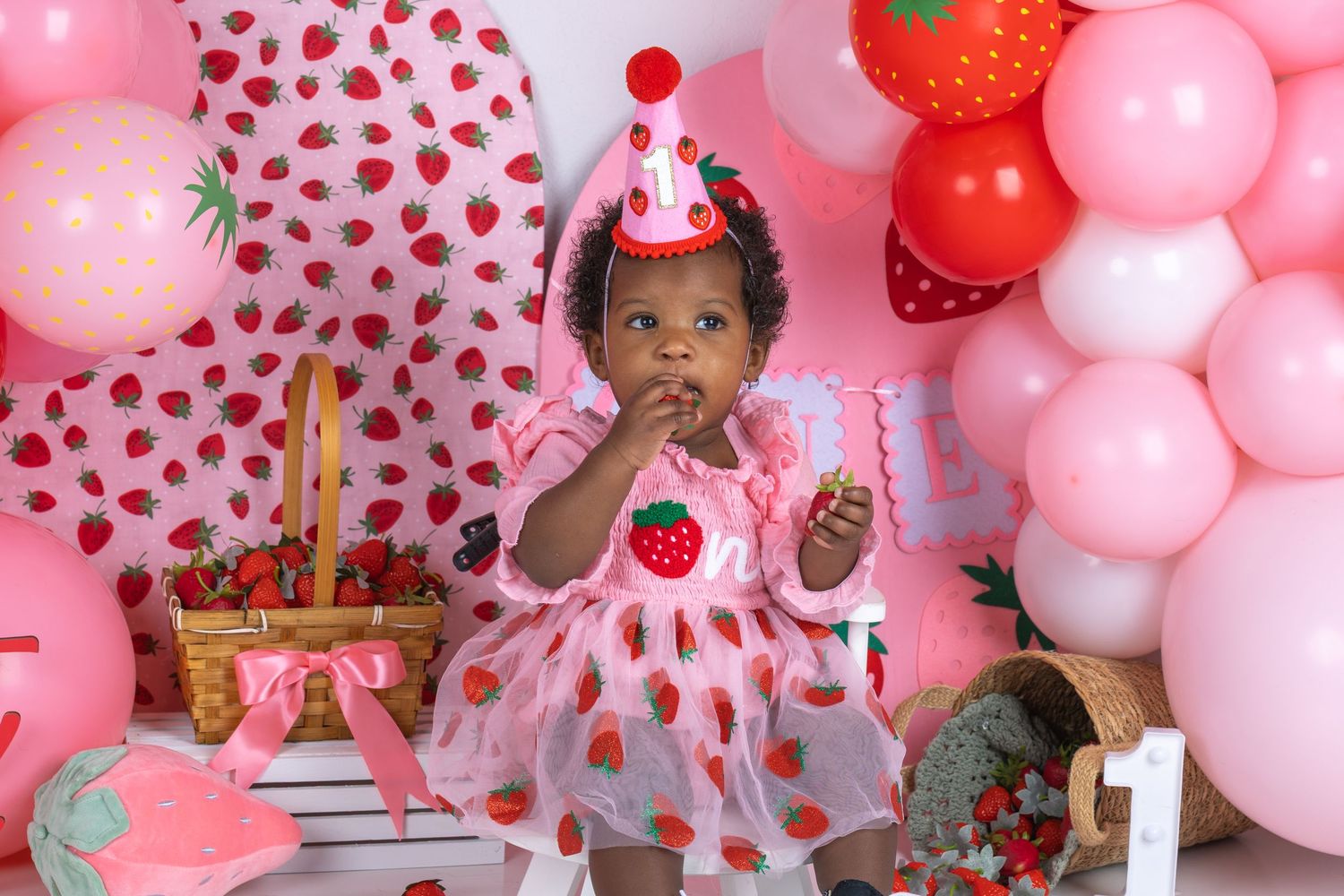 Baby girl in strawberry dress and party hat celebrates first birthday surrounded by pink balloons and strawberry decorations.