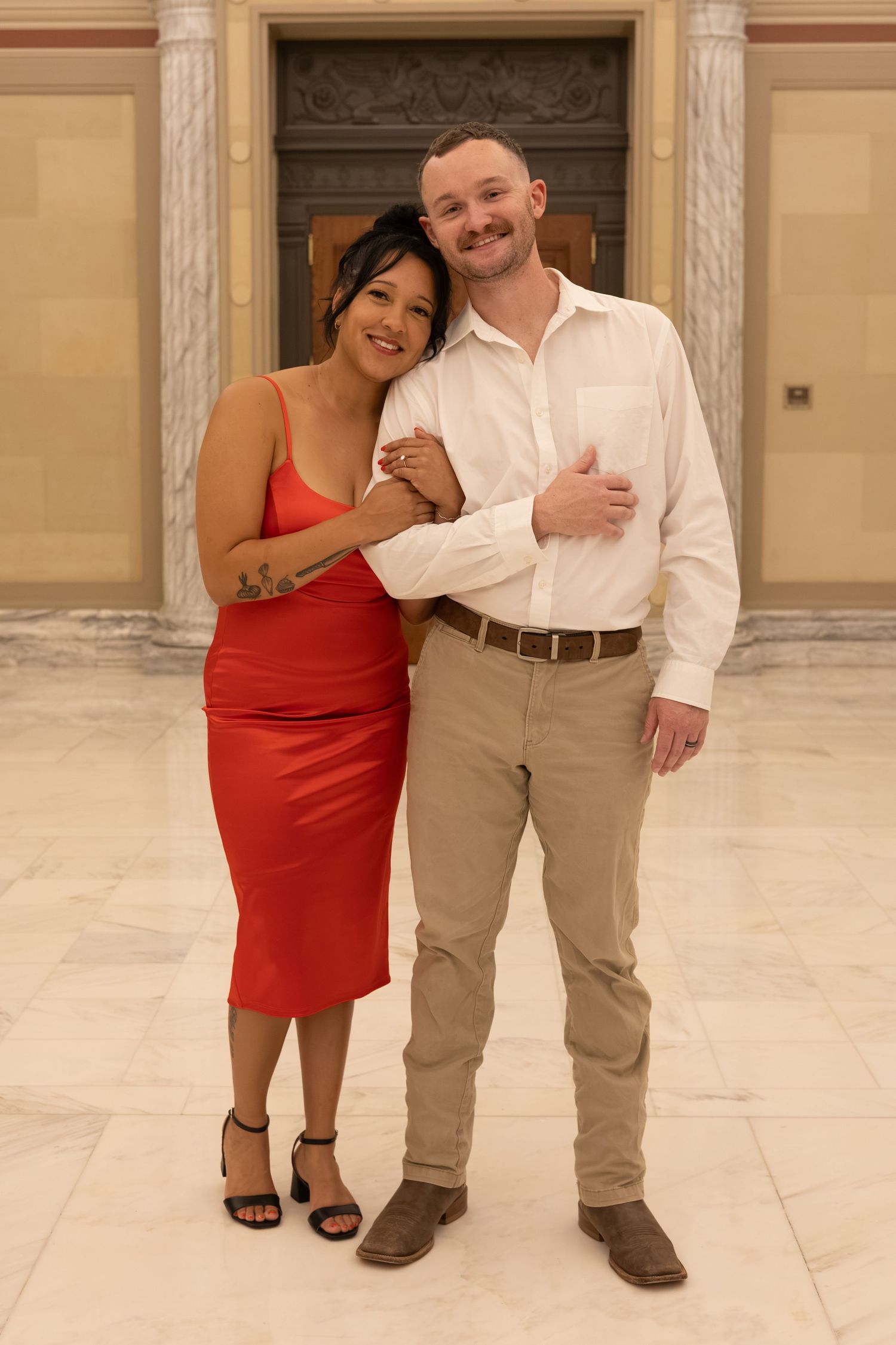 Couple posing together in elegant hall, woman in red dress and man in white shirt with khaki pants.