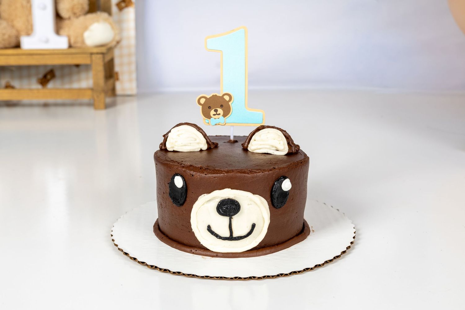 Chocolate teddy bear first birthday cake with number 1 candle and cute bear face decoration on white surface.