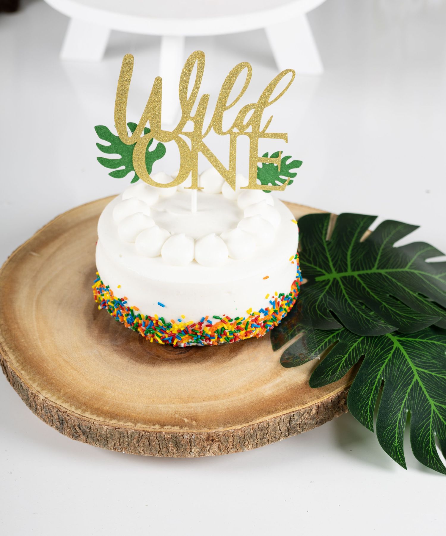 White frosted smash cake with rainbow sprinkles and gold glitter Wild One topper on rustic wood slice.