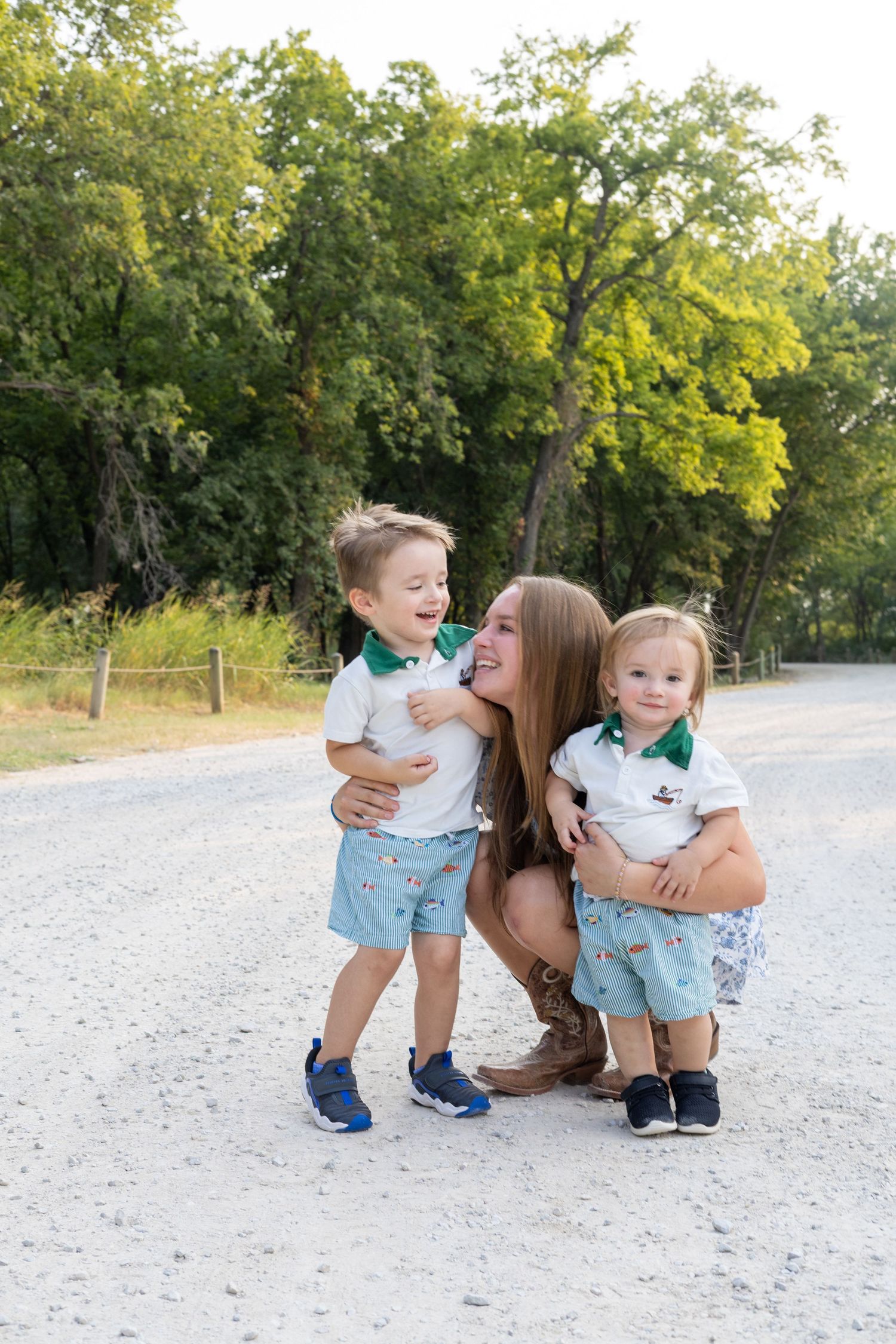 A mother crouches on a gravel path hugging her two young children in matching white and teal outfits outdoors.