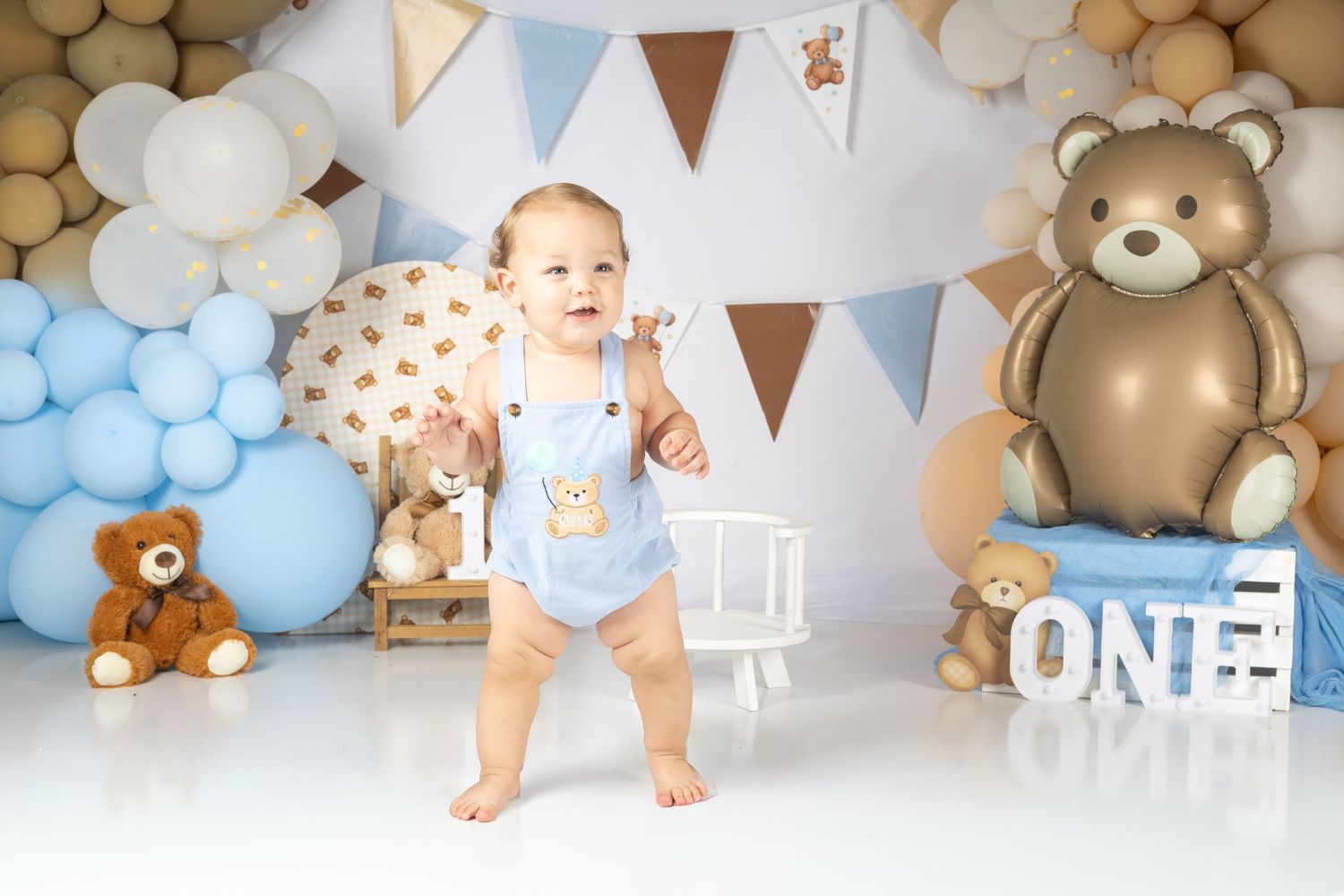 Baby's first birthday cake smash with teddy bear theme, blue balloons, bunting, and ONE sign on white backdrop.