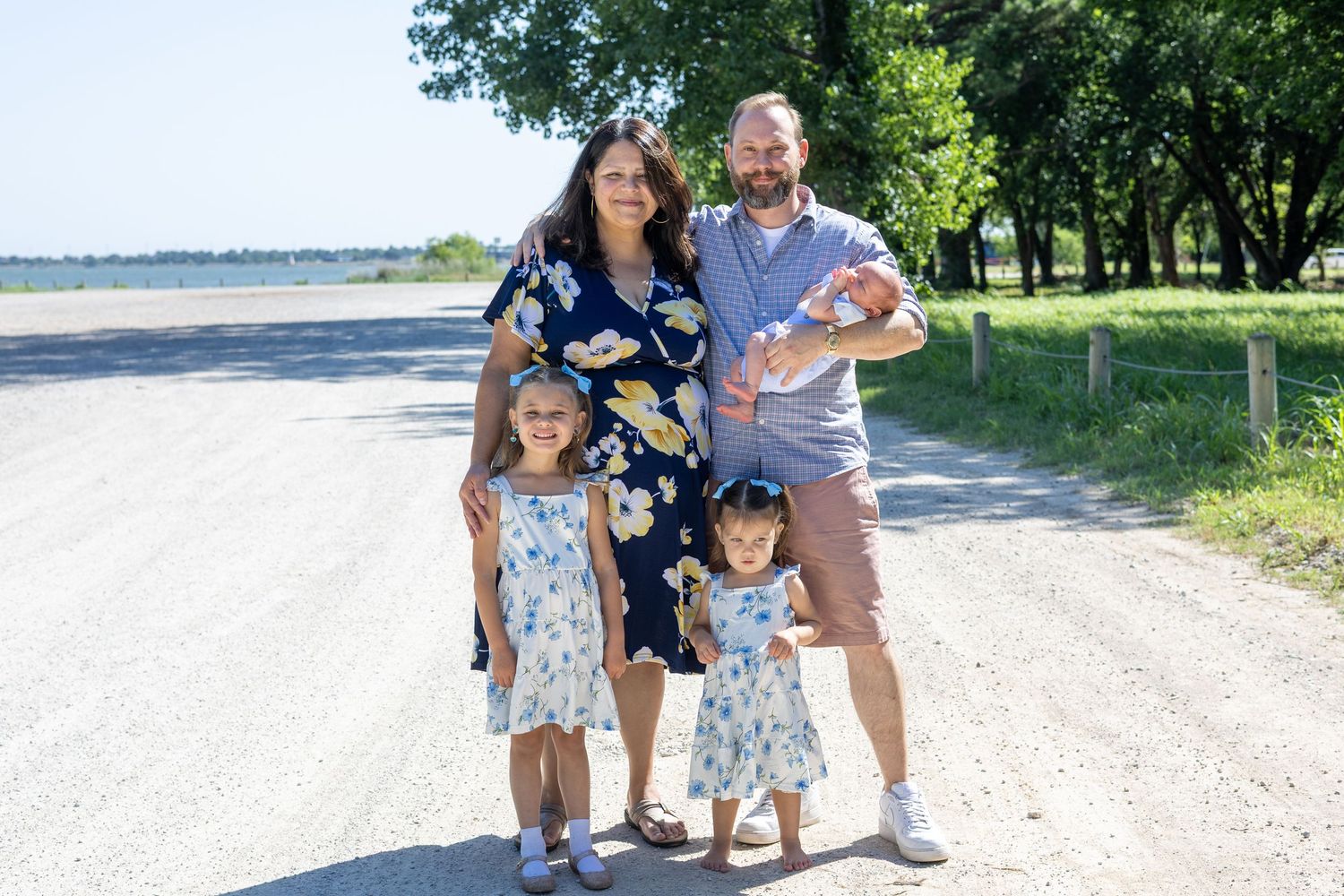 Family of five posing on a sunny rural road surrounded by green trees, parents with three young children smiling outdoors.