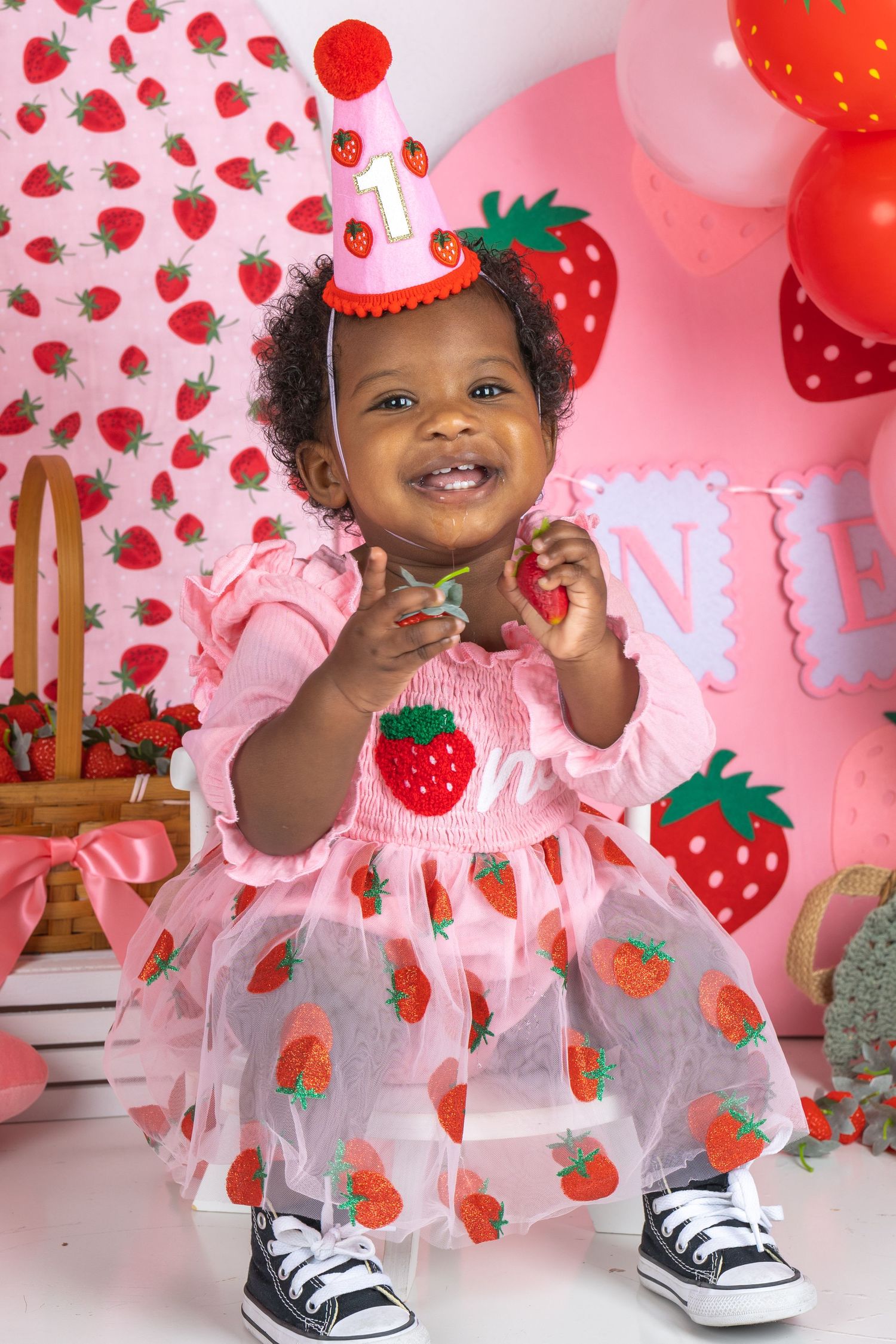 Smiling baby girl in strawberry dress and party hat celebrates her 1st birthday with colorful strawberry-themed decorations.
