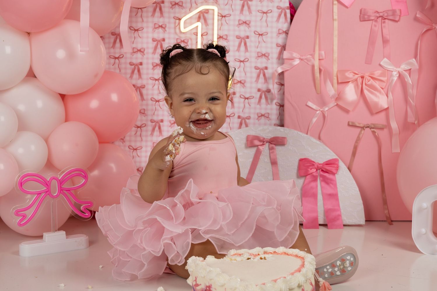 Baby girl in pink tutu celebrates first birthday smashing cake, surrounded by pink balloons and bow decorations.