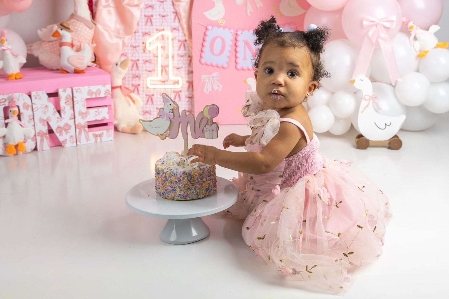 A baby girl in a pink tutu dress reaches for a sprinkle cake at her first birthday smash cake photo session.