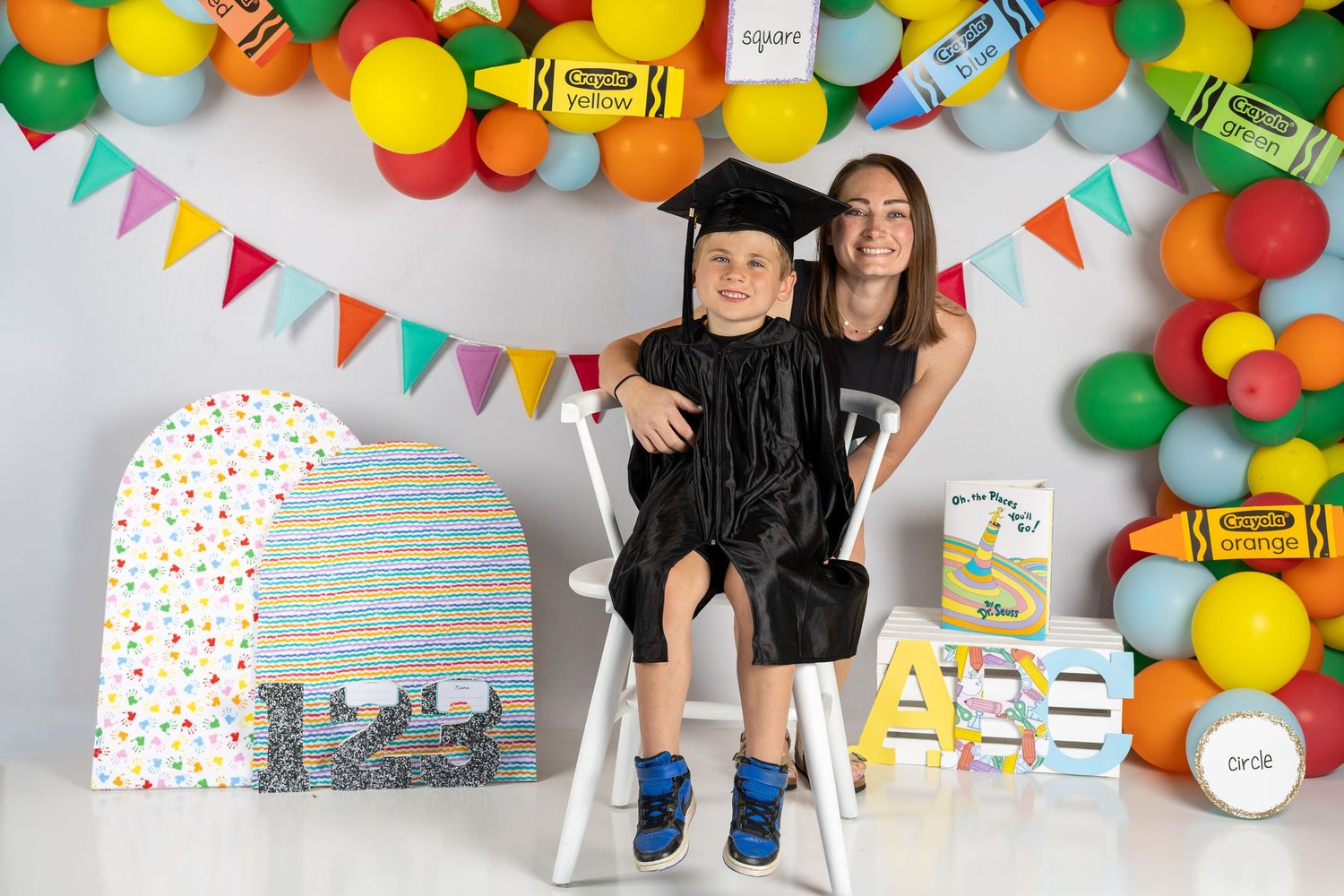 A young boy in graduation cap and gown sits with a girl amid colorful balloons and crayon decorations at a graduation party.