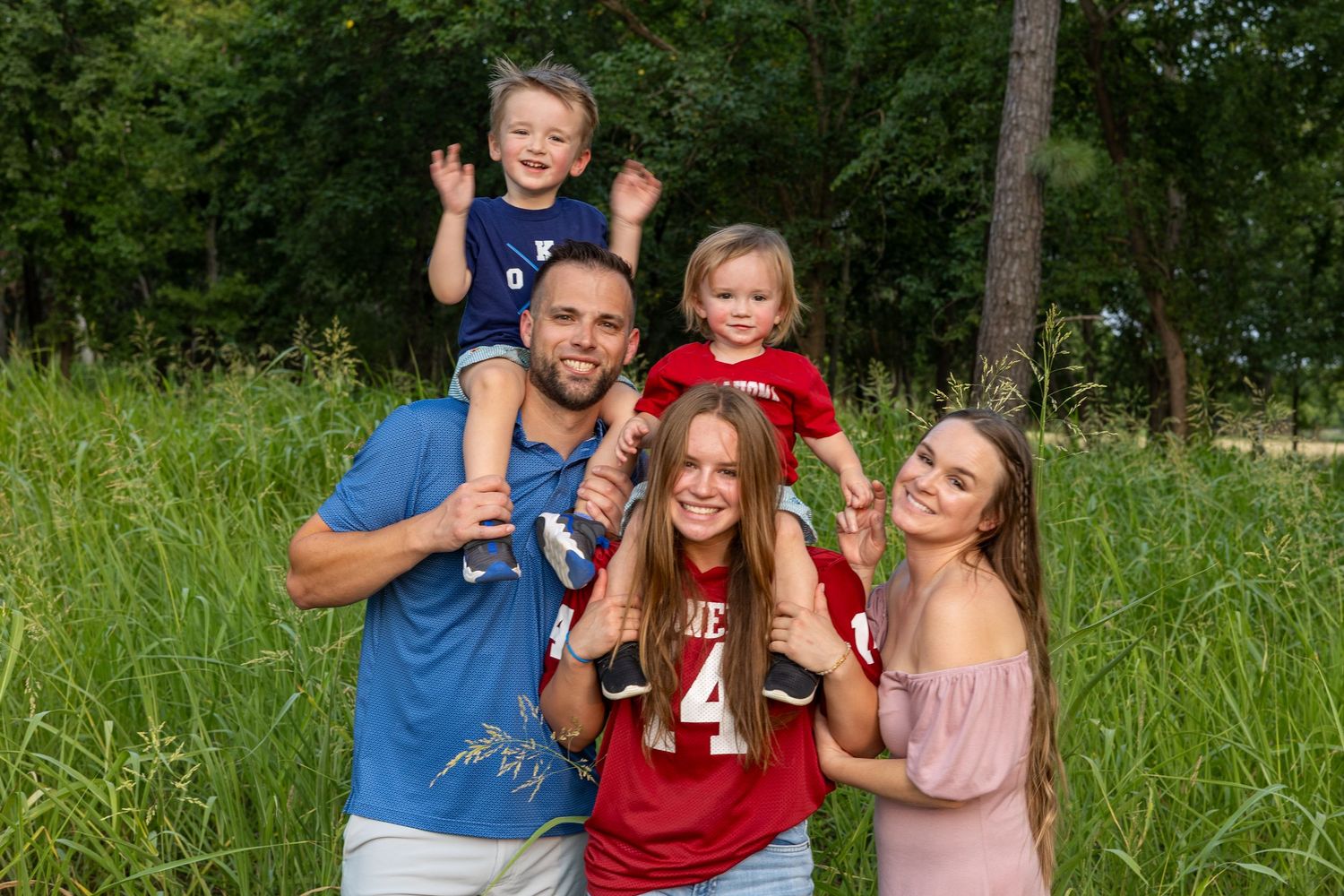 Happy family of five posing outdoors in green field, parents holding two toddlers and teenage girl smiling together.